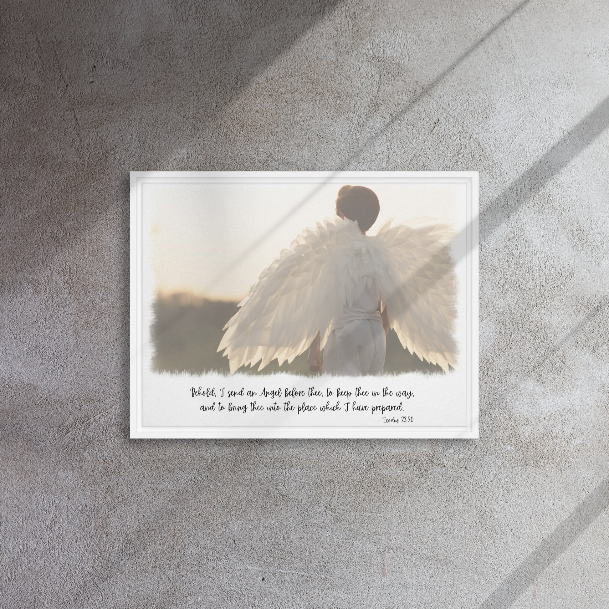 An image of the back of a realistic looking angel with spread wings looking off into the distance, with scripture below it in a script font, framed with a white frame, hangs on a gray textured wall with sunlight streaming in across it from the right.