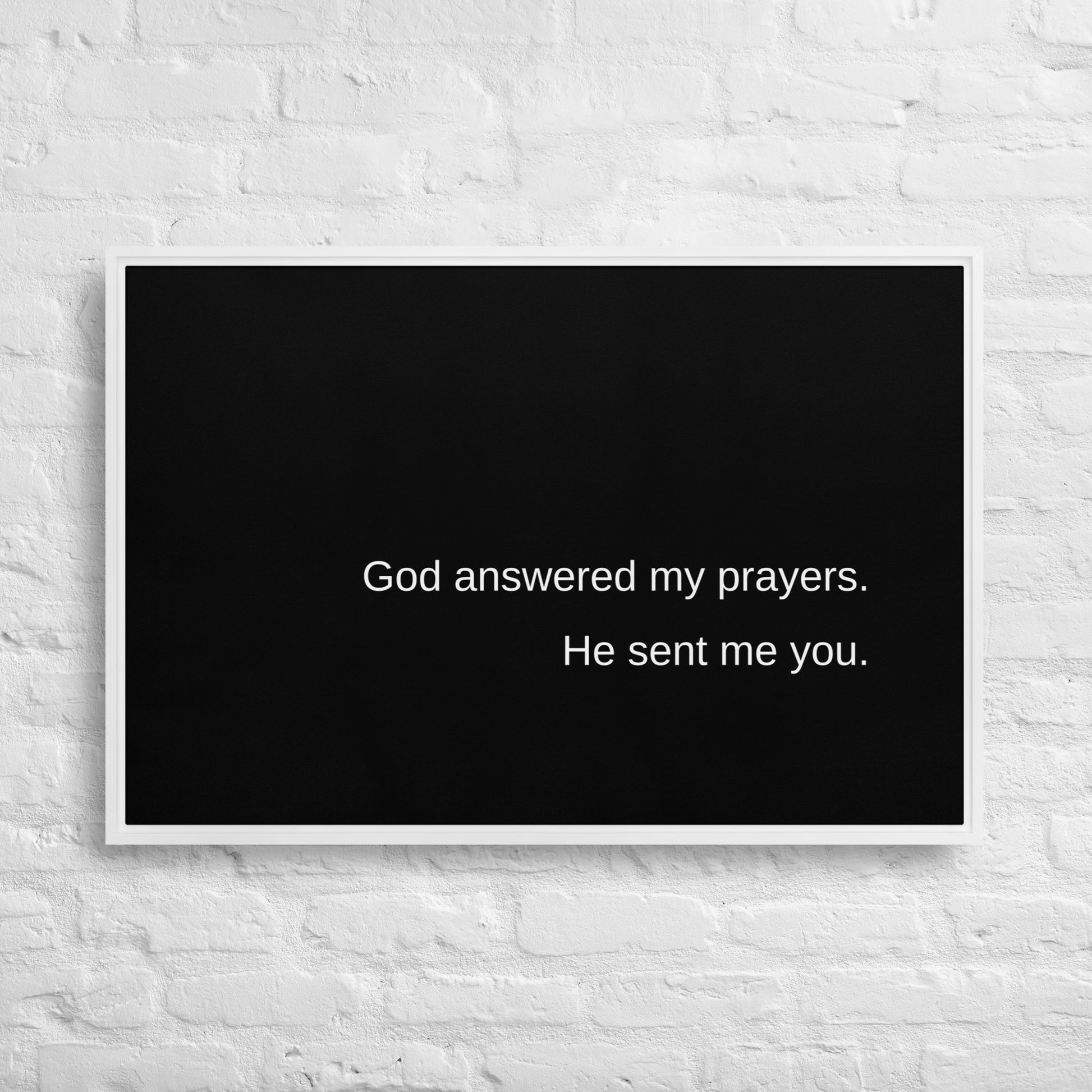 A large horizontal stretched black canvas with a white frame, and on the canvas, in white text, it says:  God answered my prayers. He sent me you. Hanging on a white brick wall.