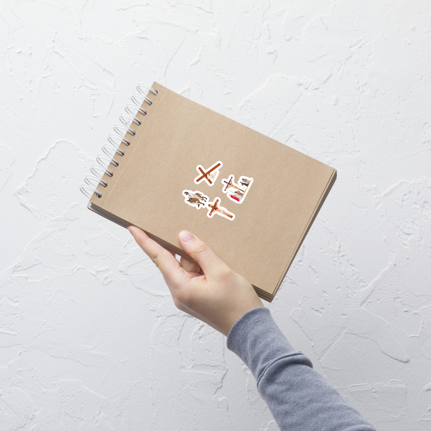 A light brown spiral bound notebook held by a woman's hand and on the notebook are four stickers of Jesus, the cross, crucifixion, and Jesus on a donkey.