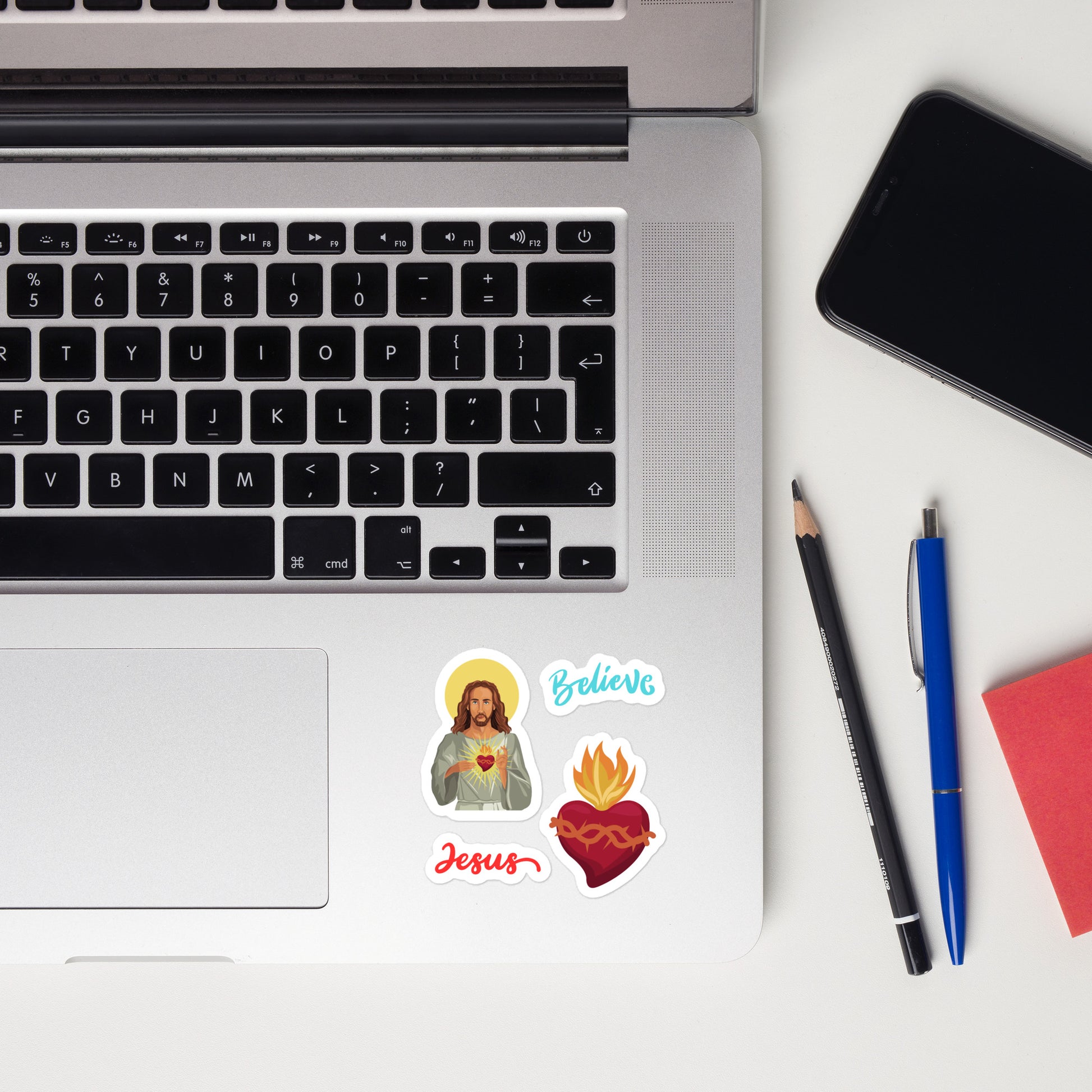 Silver laptop is open and has four stickers stuck on the lower right corner below the keyboard, one is Jesus with his Sacred heart, one is the word Jesus in red script, the word Believe in aqua script, and the fourth is just the sacred heart. Next to the laptop, there is a black cell phone, a black sharpened pencil, a blue pen, and it looks like a red pad of sticky notes.