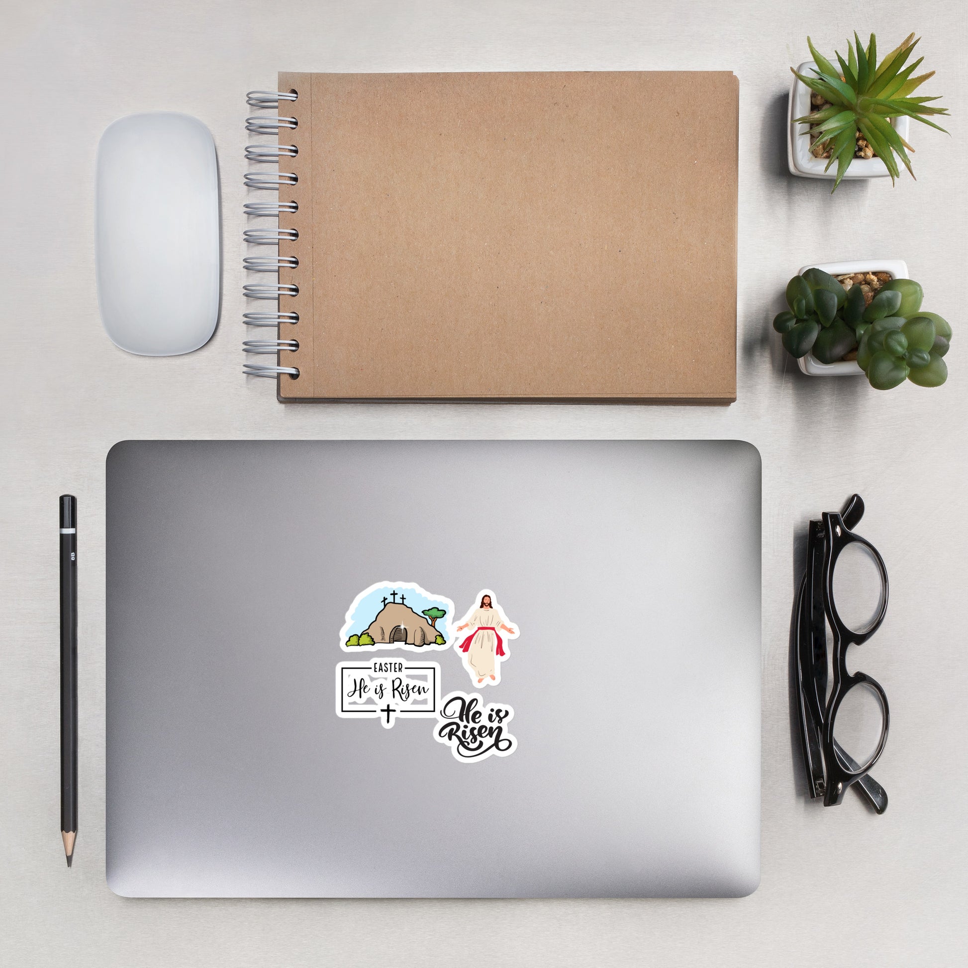 An overhead view of a silver laptop with Easter He is Risen and Jesus stickers on it, with a pair of glasses folded to the right and a pencil to the left and over it, a white computer mouse, a brown spiral bound notebook, and two succulent plants in white square pots.