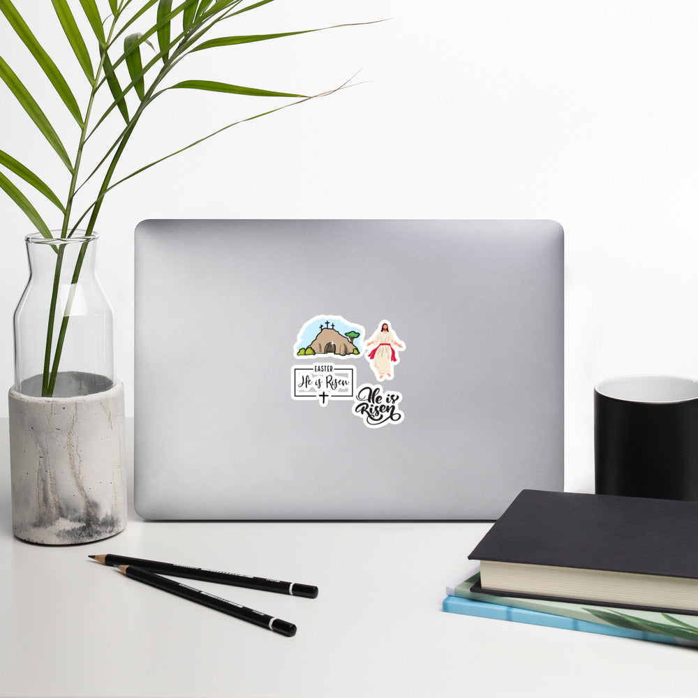 From left to right, a clear glass and white marble vase with two palm branches in it, next to a silver laptop that is somehow standing up, with four Easter He is Risen and Jesus stickers on it, then a black coffee mug, and going clockwise, a stack of three books, and two black sharpened pencils.