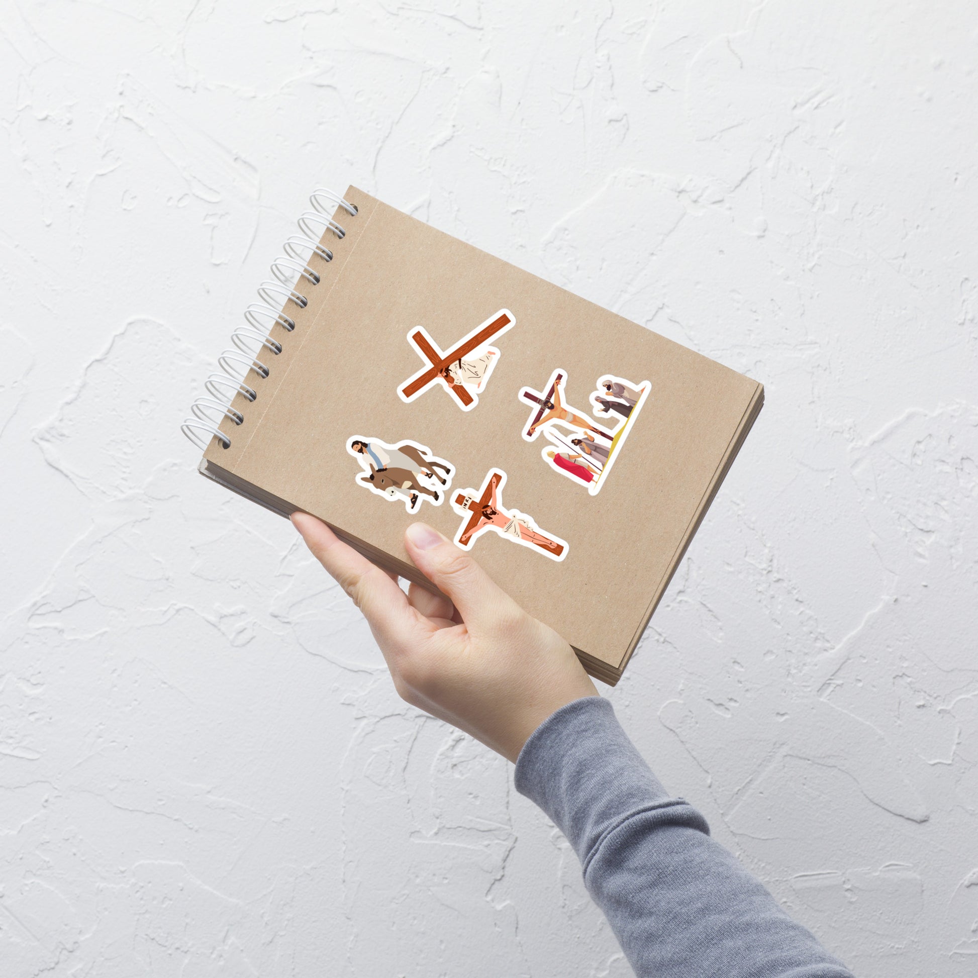 A hand holding a notebook with four stickers of Jesus and Lent time stuck on a top-spiral-bound notebook.