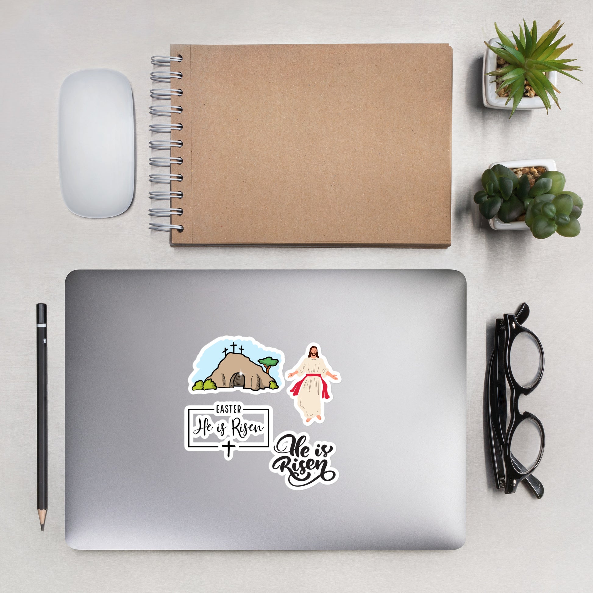 Closed laptop with Easter and Jesus stickers on it surrounded by a pencil, mouse, notebook, succulent plants in pots, and a pair of dark glasses.