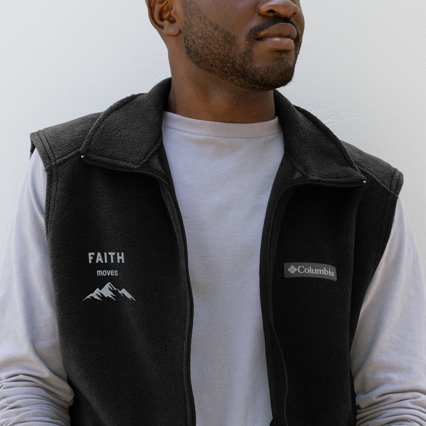 Torso and partial face view of man with facial stubble wearing light gray long sleever shirt and a black Columbia fleece vest embroidered on the right chest side with the words FAITH moves and a graphic of mountains.