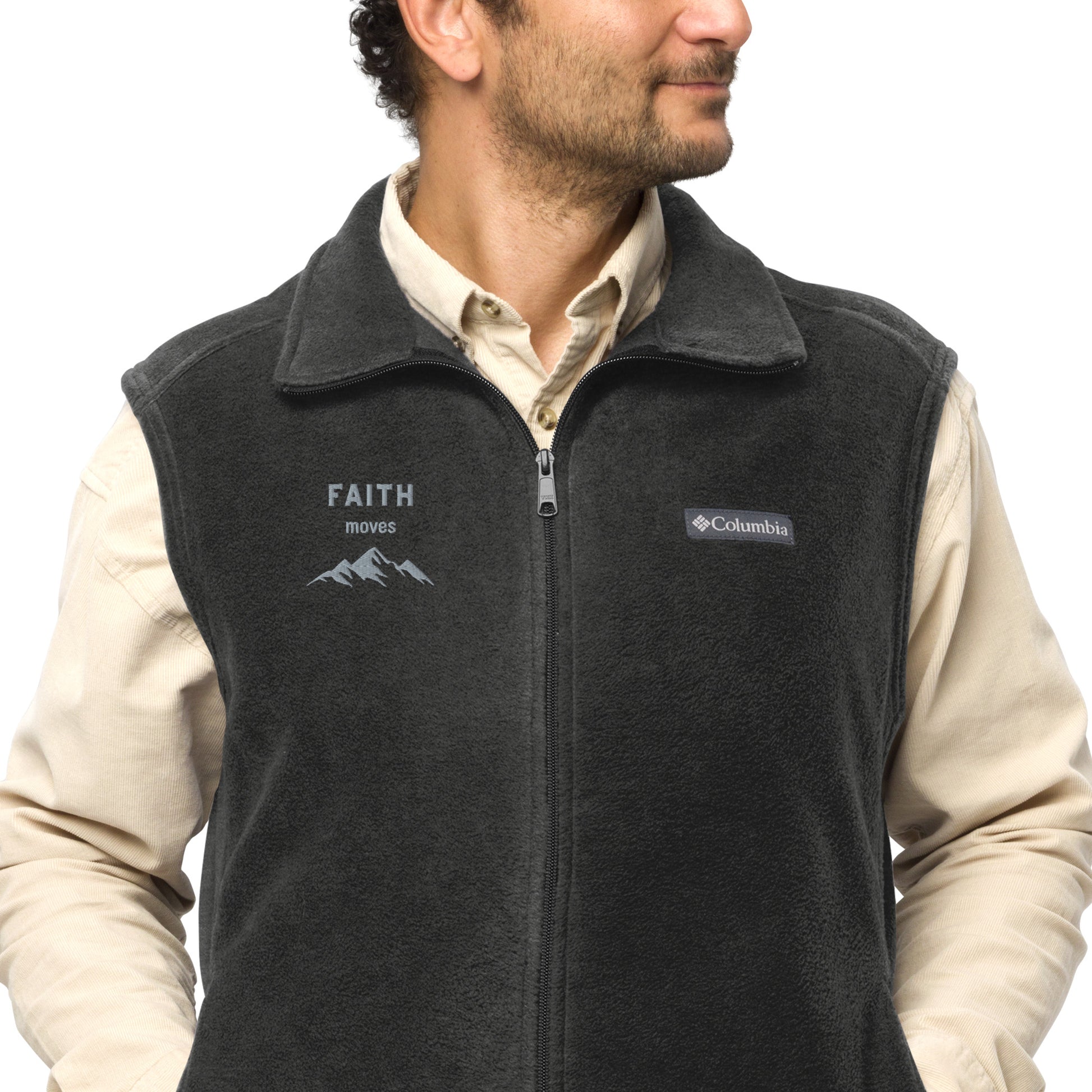 Front partial view of a man from nose to beltline with dark curly hair and short beard and mustache, wearing long-sleeve button-front cream colored shirt and a dark gray or graphite colored Columbia sleeveless vest, zipped up, with the words FAITH moves and a mountain graphic embroidered on the right front chest area.