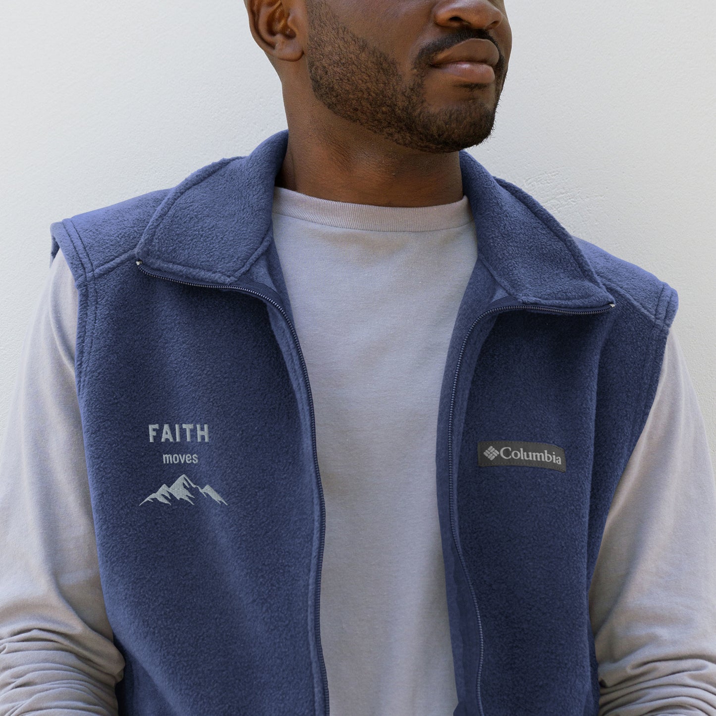 Photo of mid-section of man with facial stubble wearing long sleeve gray shirt with sleeveless Columbia fleece vest over top, embroidered on the right chest area with the words FAITH moves and a graphic of mountain peaks.