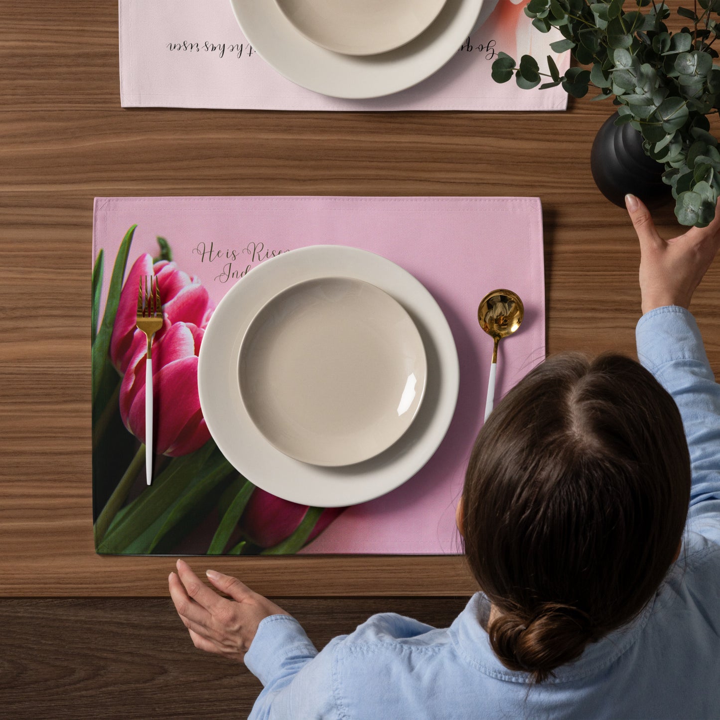 Pink fabric placemat with tulips on it on dining table with dishes and silverware, woman, and plant taken from above.