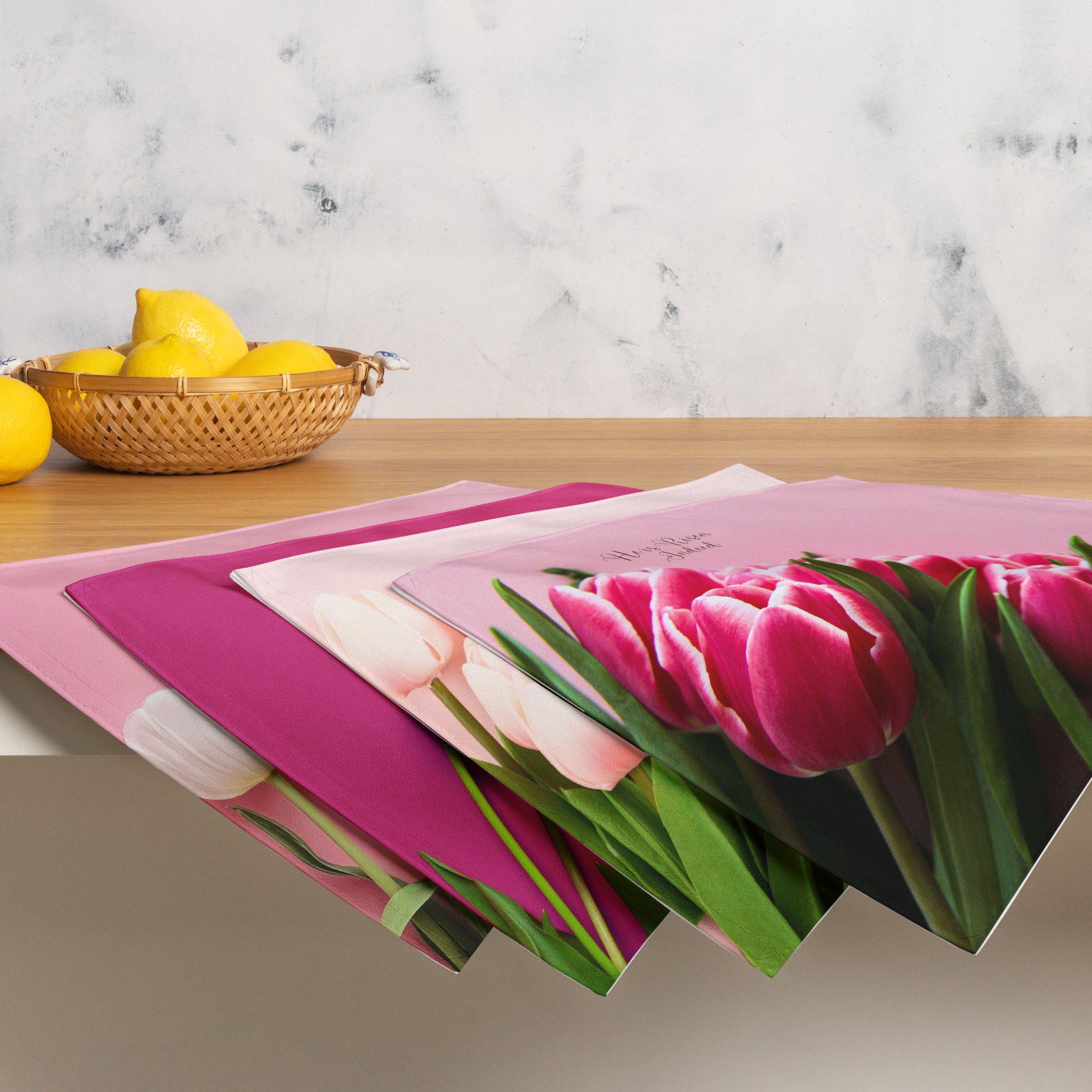 Four different pink fabric placemats with tulips on them hanging over the edge of a counter and a bowl of yellow lemons on counter