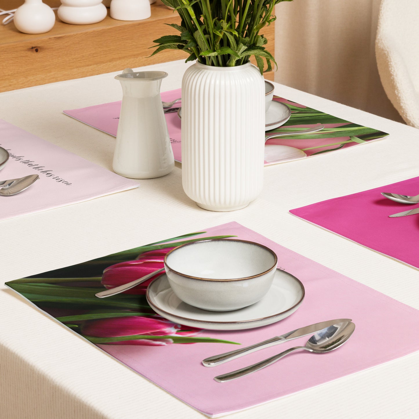 Pink fabric tulip placemats on white table with dishes and silverware set at place settings and vase in the middle.