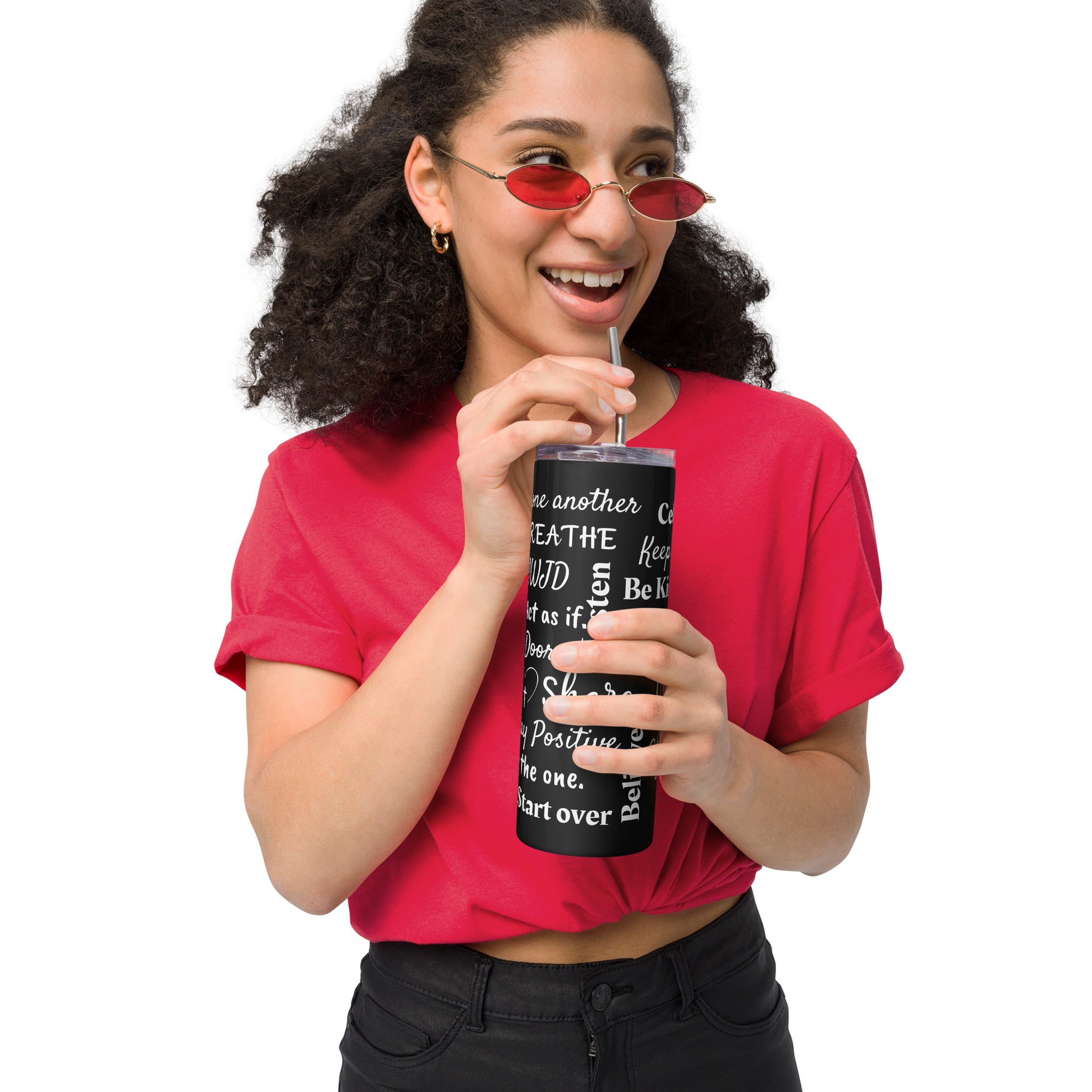 A woman with dark curly hair and sunglasses is wearing black jeans and a red short-sleeved shirt, holding a black stainless steel tumbler with a clear cover and metal straw that has white words all over it.