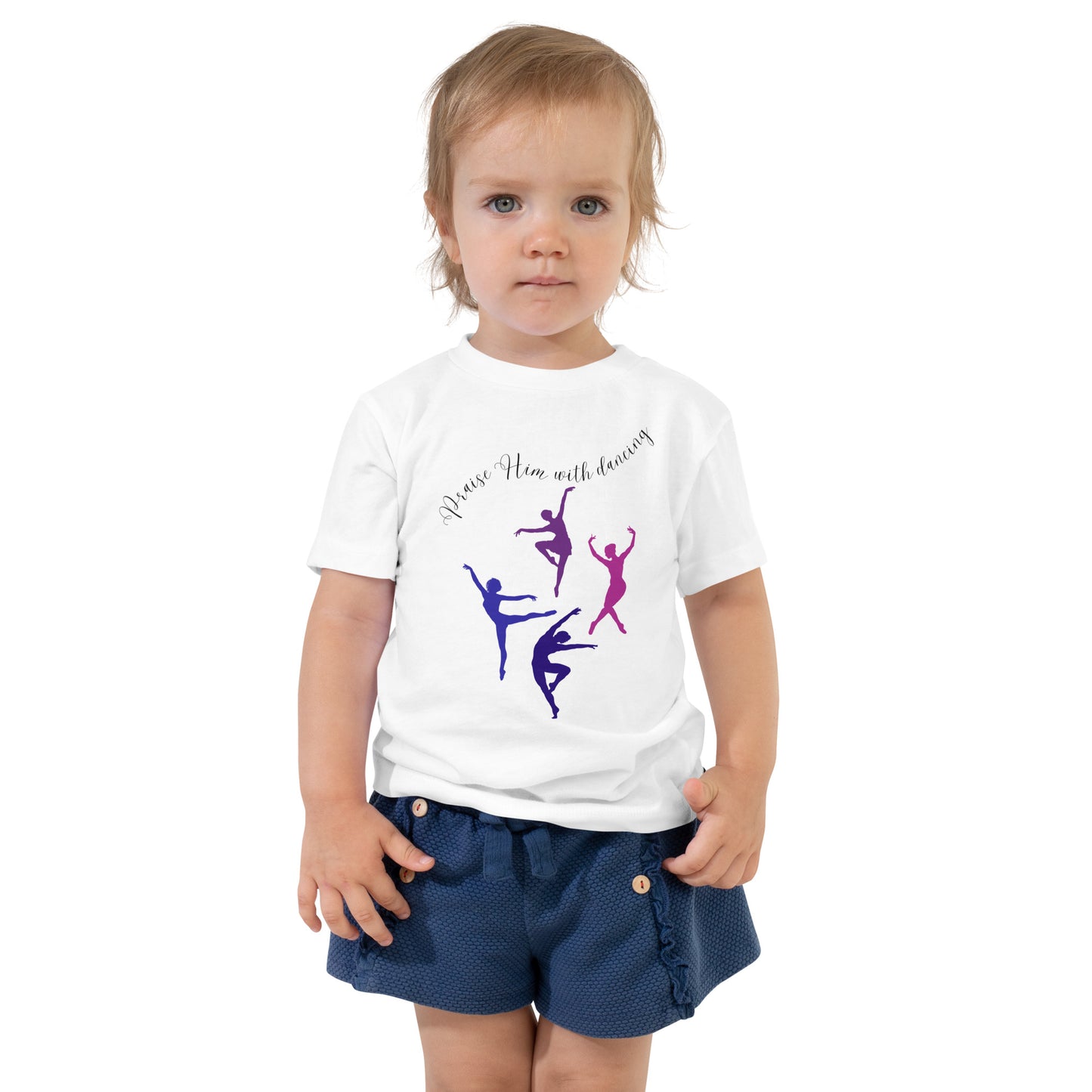 A serious-looking toddler girl with short hair and big blue eyes is wearing navy shorts or a skort with buttons on it and a short-sleeved white t-shirt with four dancers in different dance poses on the front in different colors and the words:  Praise Him with dancing.