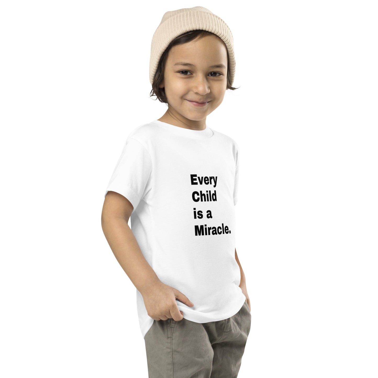 A smiling toddler wearing gray pants and a knit hat and a short-sleeve white t-shirt that says "Every Child is a Miracle." in black text.