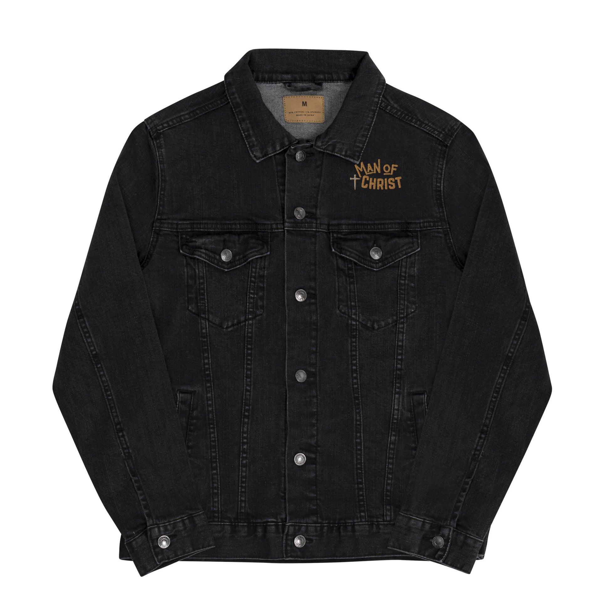 A black denim jacket on a solid white background and the denim jacket has silver metal buttons down the front and on the cuffs and the two front pocket flaps, stitching down the front, and an embroidered design on the left lapel area above the pocket that is a cross made of nails in gray and then the words Man of Christ embroidered in gold thread.