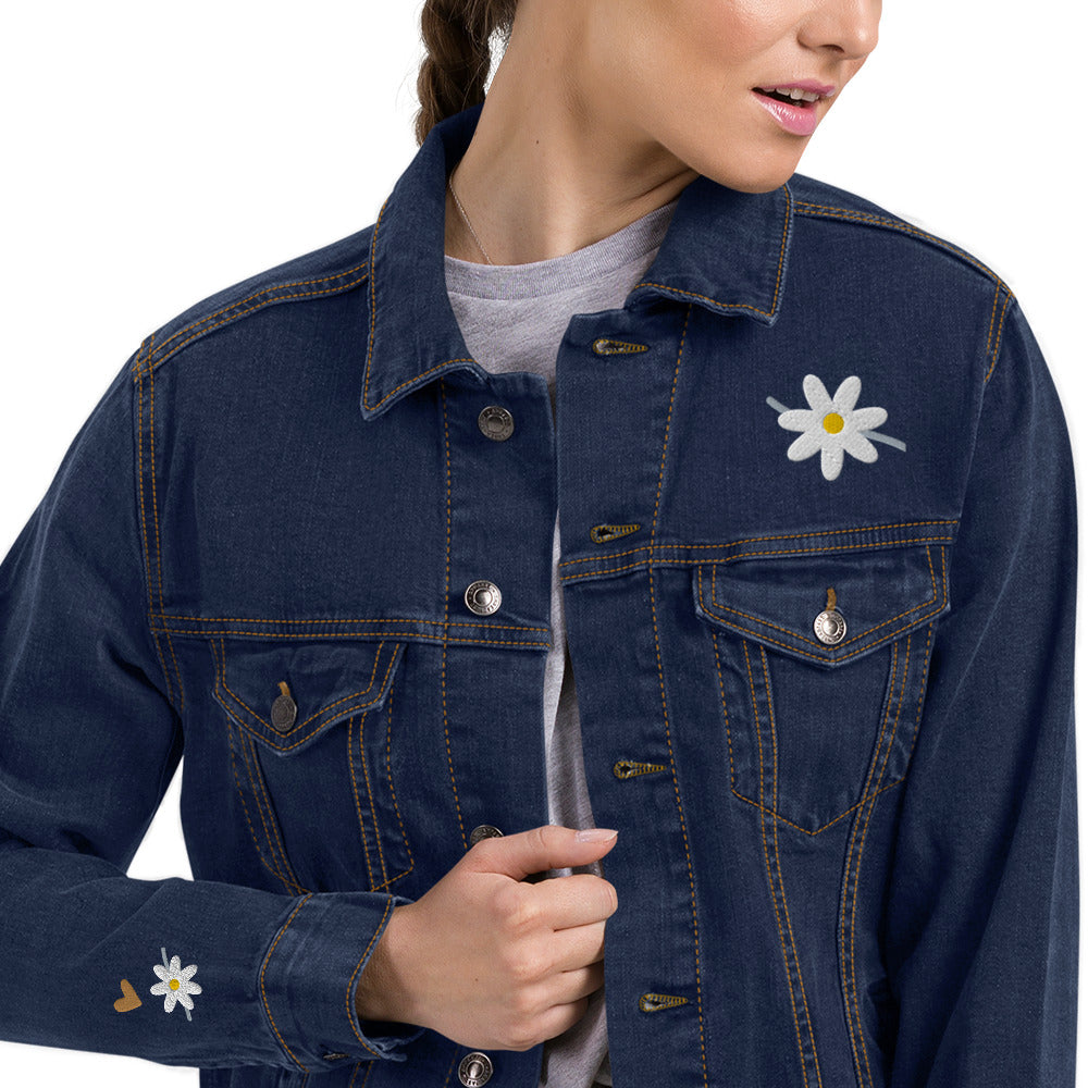 Woman with hair in braid is wearing a gray t-shirt over which she wears a blue denim jean jacket embroidered with a white daisy on the front left lapel and a heart and daisy on the lower arm area.