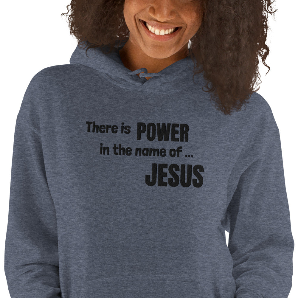 A view of a dark curly-haired smiling woman from just below the eyes to about her hips. She is wearing a heather gray hoodie and the front is embroidered in black stitching that says There is POWER in the name of JESUS