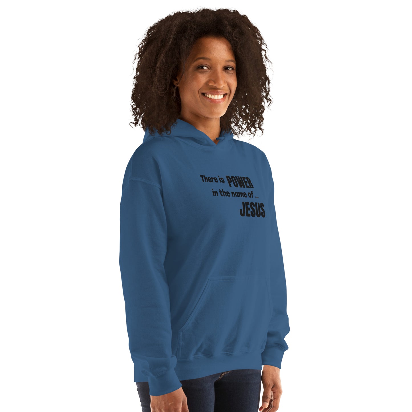 A three-quarter length photo of a woman standing about 45 degrees from camera facing, with shoulder length dark curly hair, smiling  with hands down at her sides, wearing dark jeans and a blue sweatshirt that has dark embroidered words on the front:  There is POWER in the name of JESUS.