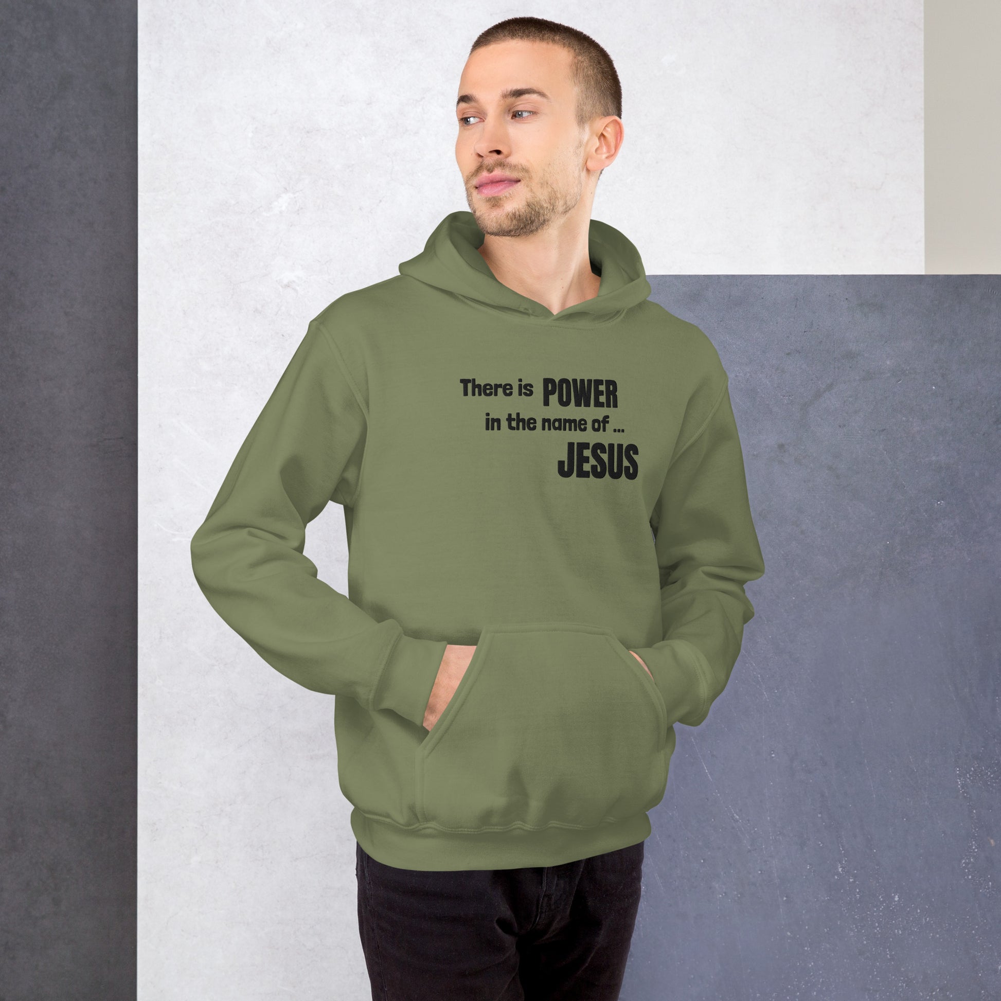 A three-quarter length photo of a man standing in front of a neutral colored block background wearing black pants and an olive or army green colored hooded sweatshirt or hoodie with both hands in the front pouch pocket and the front of the shirt is embroidered to say There is POWER in the name of JESUS.