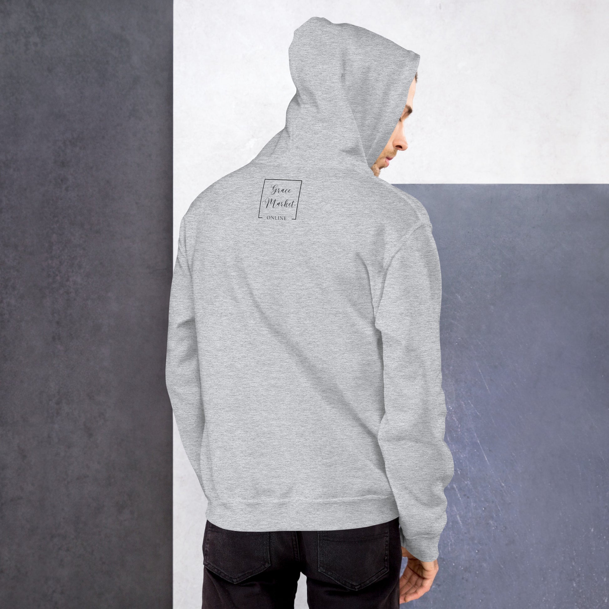 A man in a light gray hoodie facing a white and gray color blocked wall. He is wearing black jeans and has the hood of the sweatshirt up almost obscuring his head except for a small part of his face is showing, and the sweatshirt is imprinted on the back with the words Grace Market Online.