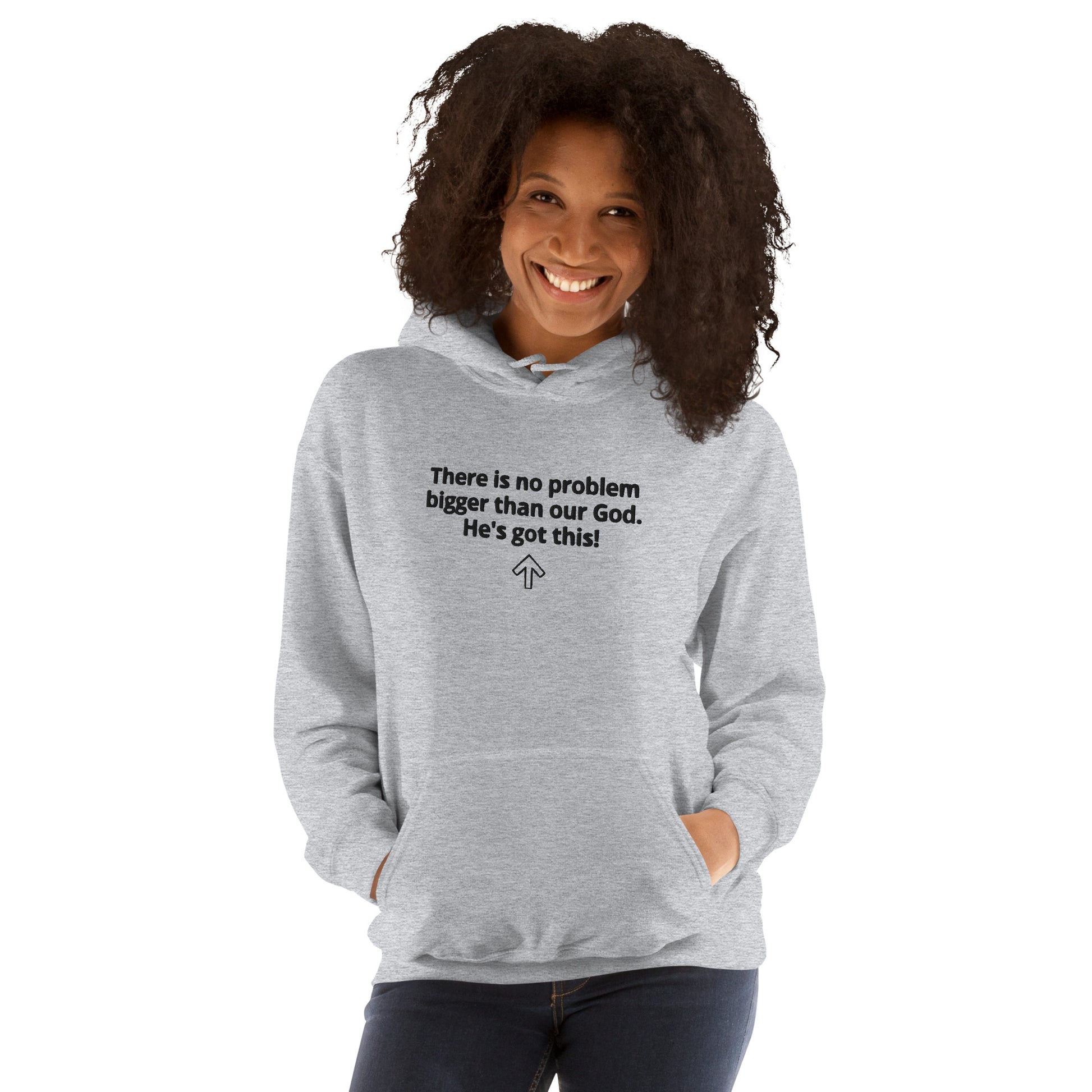 A three-quarter length view of a woman wearing jeans and a light gray hooded sweatshirt with both of her hands tucked into the front pouch pocket and the sweatshirt says "There is no problem bigger than our God. He's got this!" and an upward arrow symbol is below it and she is smiling.