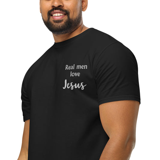 View of man's lower face with dark bear and mustache and muscular arms wearing black short-sleeved t-shirt with white embroidered in the left chest pocket area that says:  Real men love Jesus.