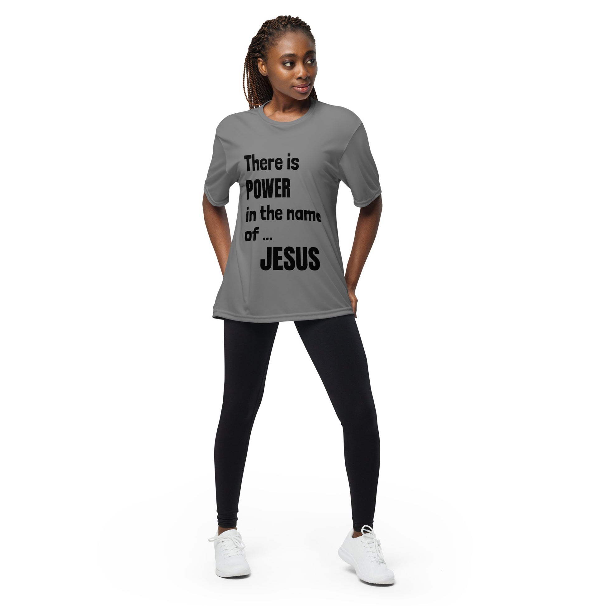 Confident woman with dark hair in cornrows is wearing white shoes, black leggings, and a light gray t-shirt that says There is POWER in the name of JESUS and she has her hands sort of near her thighs behind her. 