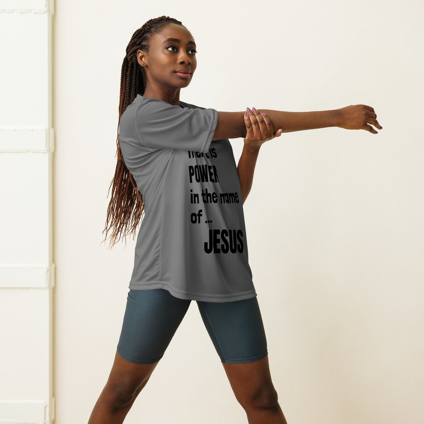 Three-quarter length photo of a woman with long hair in cornrows wearing blue bicycle shorts and a gray t-shirt with words on it stretching out her arms, and the words that are visible say Power in the name of JESUS.