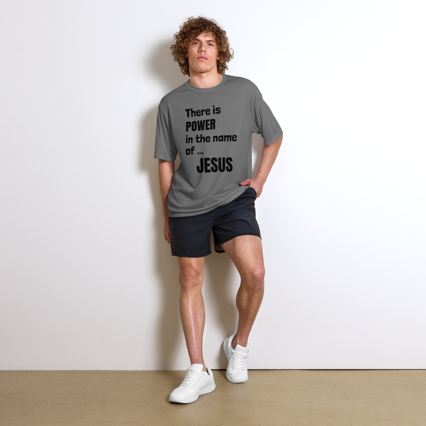 Full length photo of a young man with curly reddish hair leaning against a white wall with one hand partially in the pocked of his navy shorts. He is also wearing white sneakers and a gray short-sleeved t-shirt that proclaims:  There is POWER in the name of JESUS in black letters.