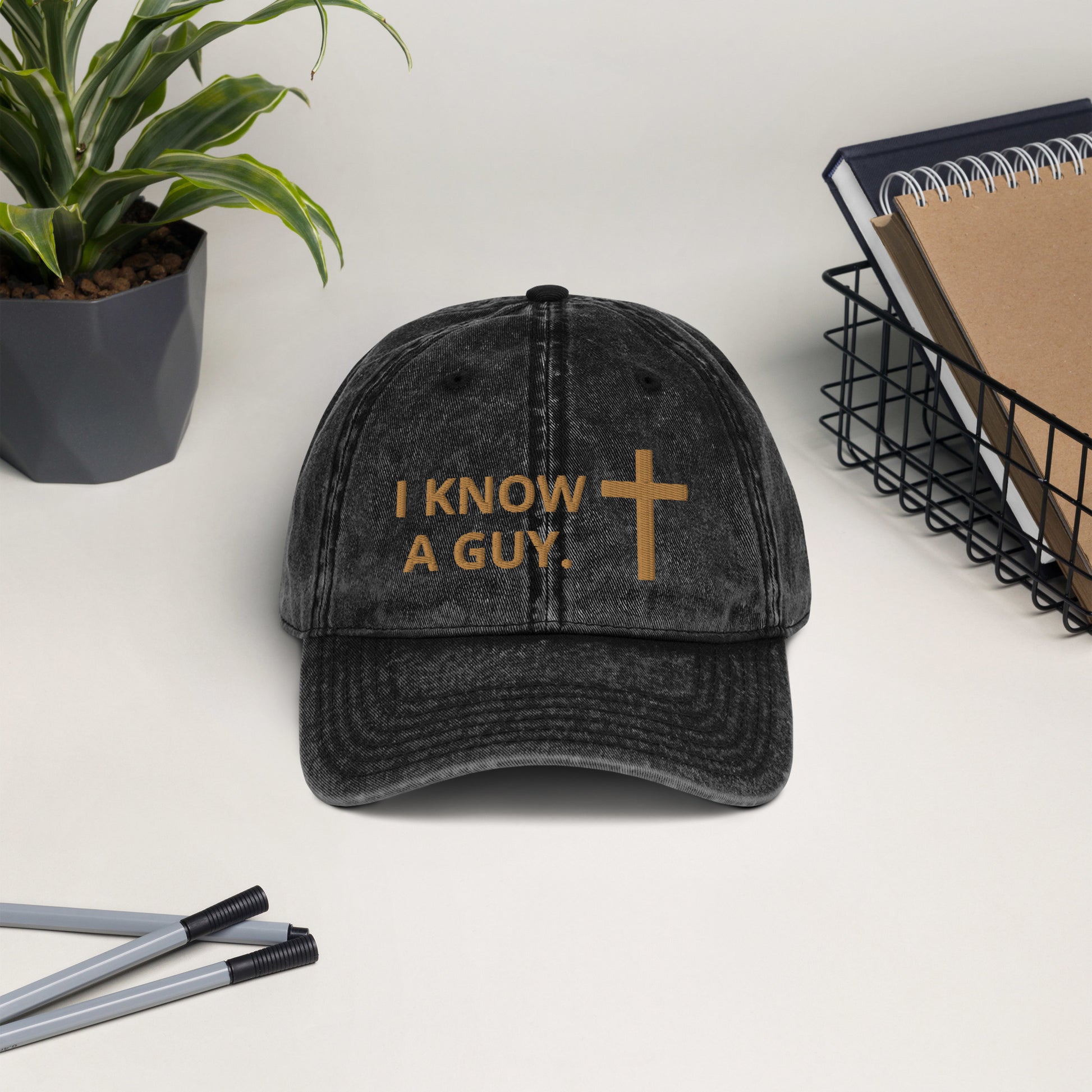 A white background with pens, a potted green plant, a wire basket with a book and notebook in it, and in the center, a vintage style washed out black cap embroidered in gold with I KNOW A GUY. and a cross.