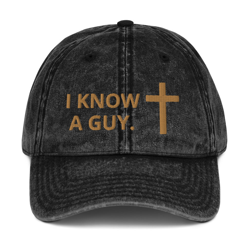 Black washed-out looking cap embroidered with the phrase "I know a guy" in gold and a gold cross on a plain white background.