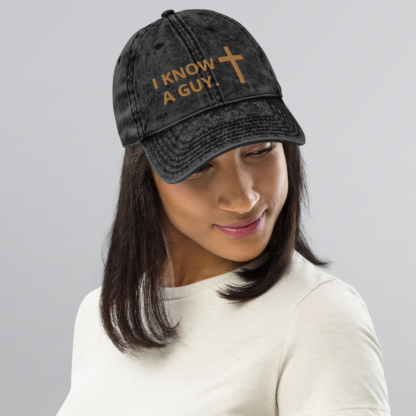 Smiling dark-haired woman with chin tilted down to show the embroidery on the washed out style black cap she's wearing that has "I know a guy" and a cross embroidered on it in gold letters.