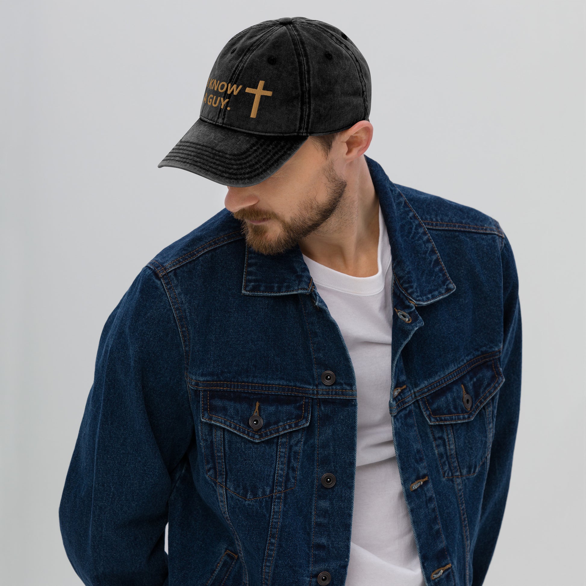 A young man with mustache and beard, wearing a white t-shirt and blue jean jacket and a black stone-washed kind of baseball cap with gold words and a gold cross.