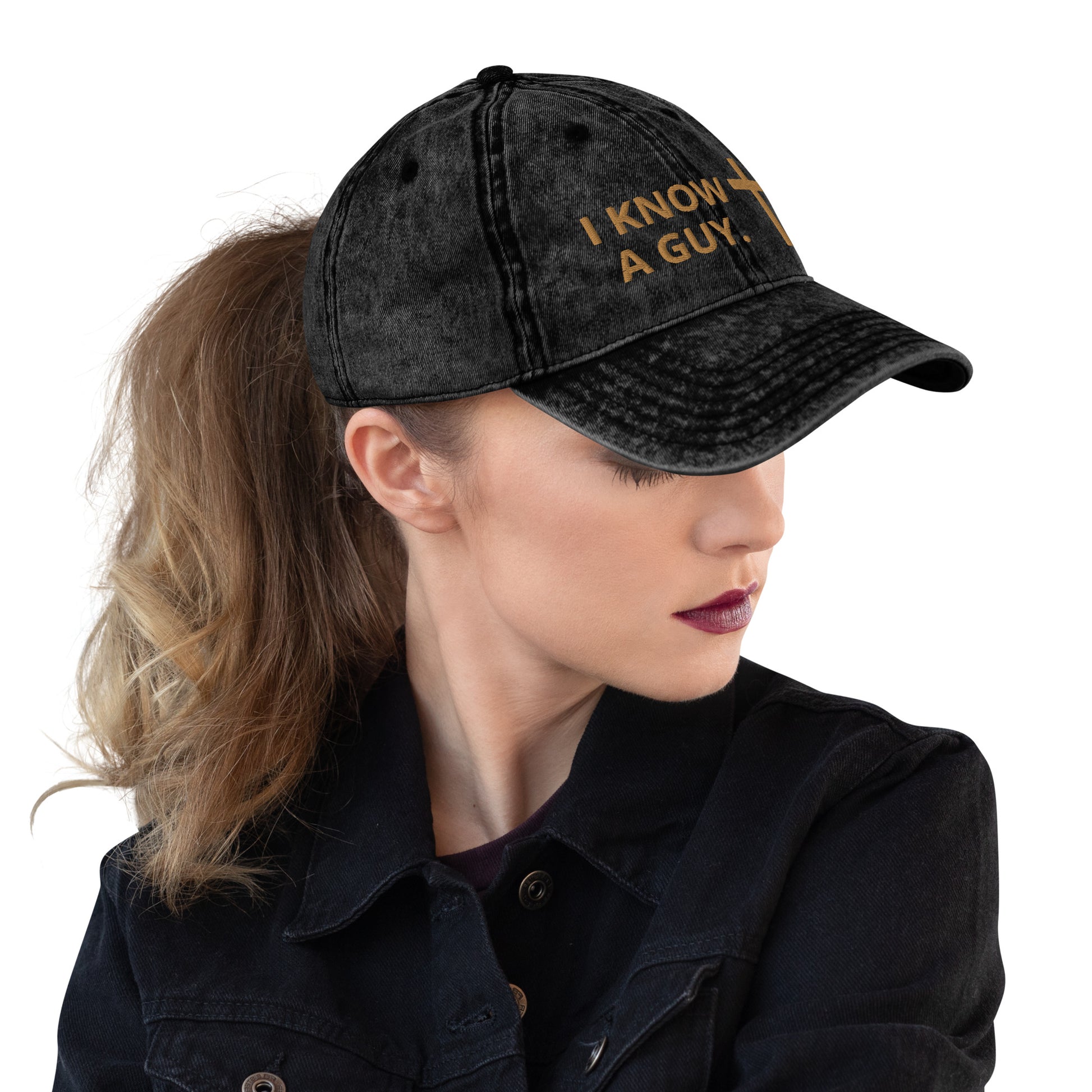 Blondish-brown haired woman with hair in pony tail coming out of the back of a black cap with gold embroidered lettering and a cross. It's says I KNOW A GUY.