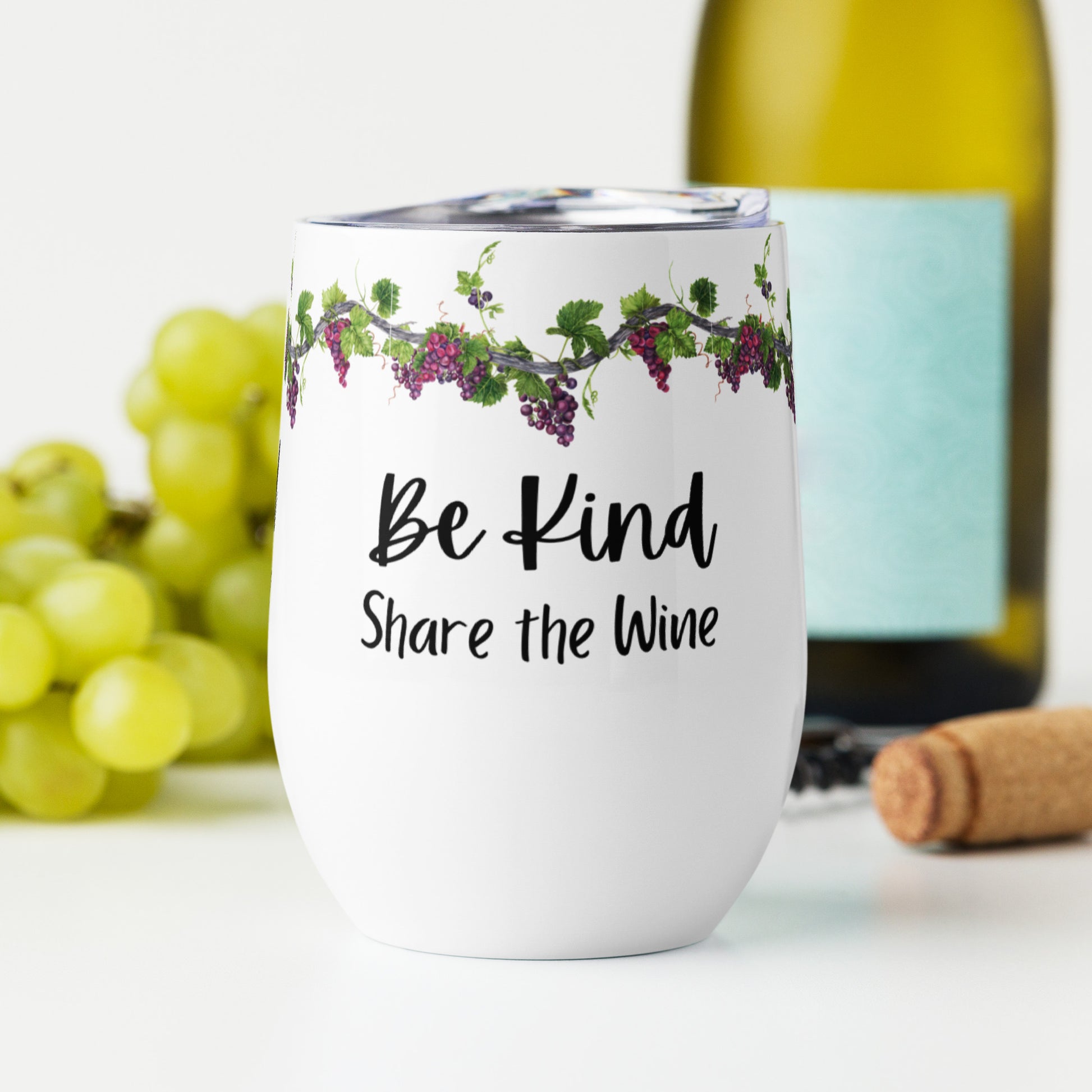 Stainless steel wine tumbler with grapevines with purple grapes running along the top and the words "Be Kind Share the Wine" on a white background, sitting on a white table with green grapes, a bottle of wine, and a corkscrew behind it.