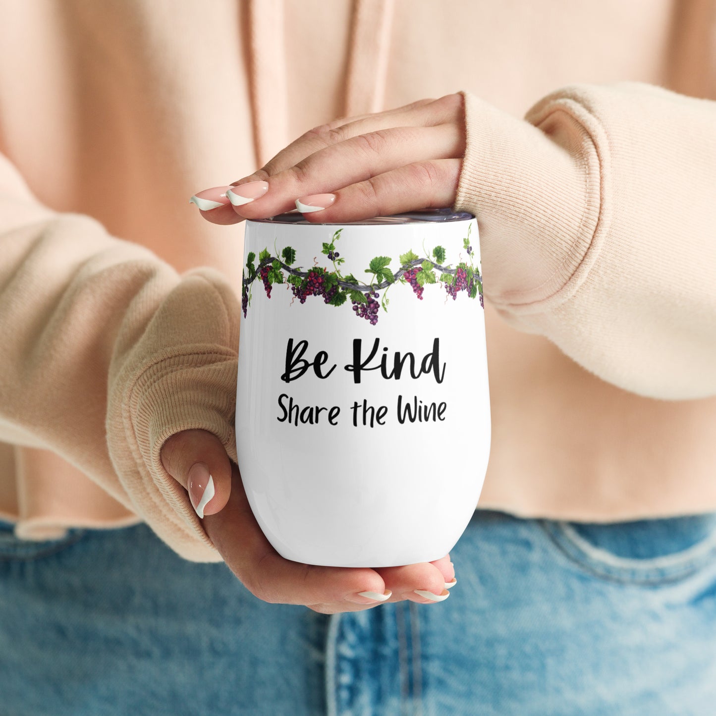 The midsection of a woman wearing a peach colored sweatshirt and bluejeans and in her manicured hands, she is holding a stainless steel wine tumbler that is white with purple grapes on grapevines and below it the words "Be Kind Share the Wine" and one hand is resting on top.