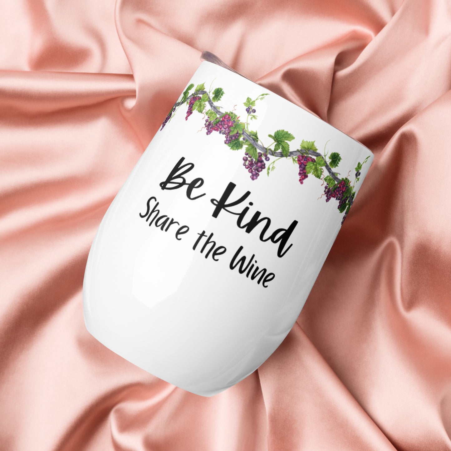 Silky satin fabric crunched up and a stainless steel wine tumbler is lying on the fabric; the sides were white with the words "Be Kind Share the Wine" in two different fancy fonts and along the top is a border of grapevines with green leaves and purple grapes on it.