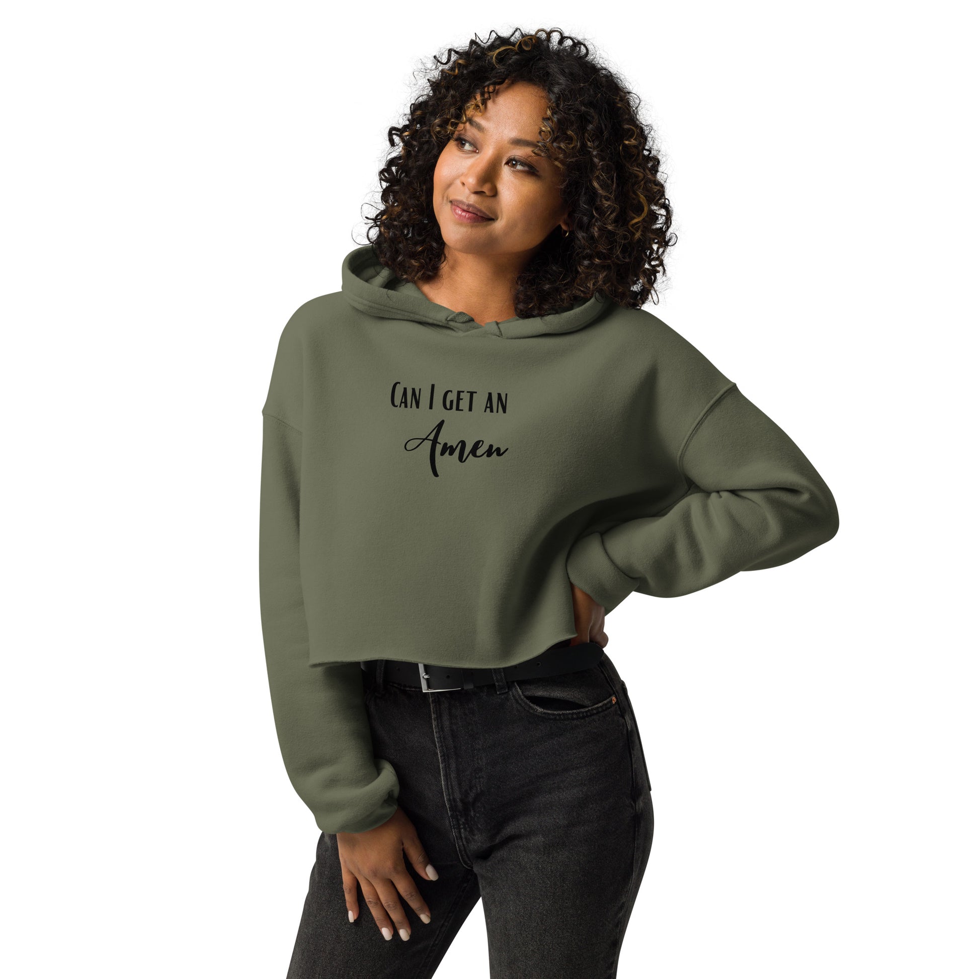 Curly haired woman is smiling and looking off camera, with one hand on her hip and the other on her thigh, and she is wearing black jeans with a belt with silver buckle and a military green, long-sleeved, hooded crop top with the words Can I Get an Amen on the front.