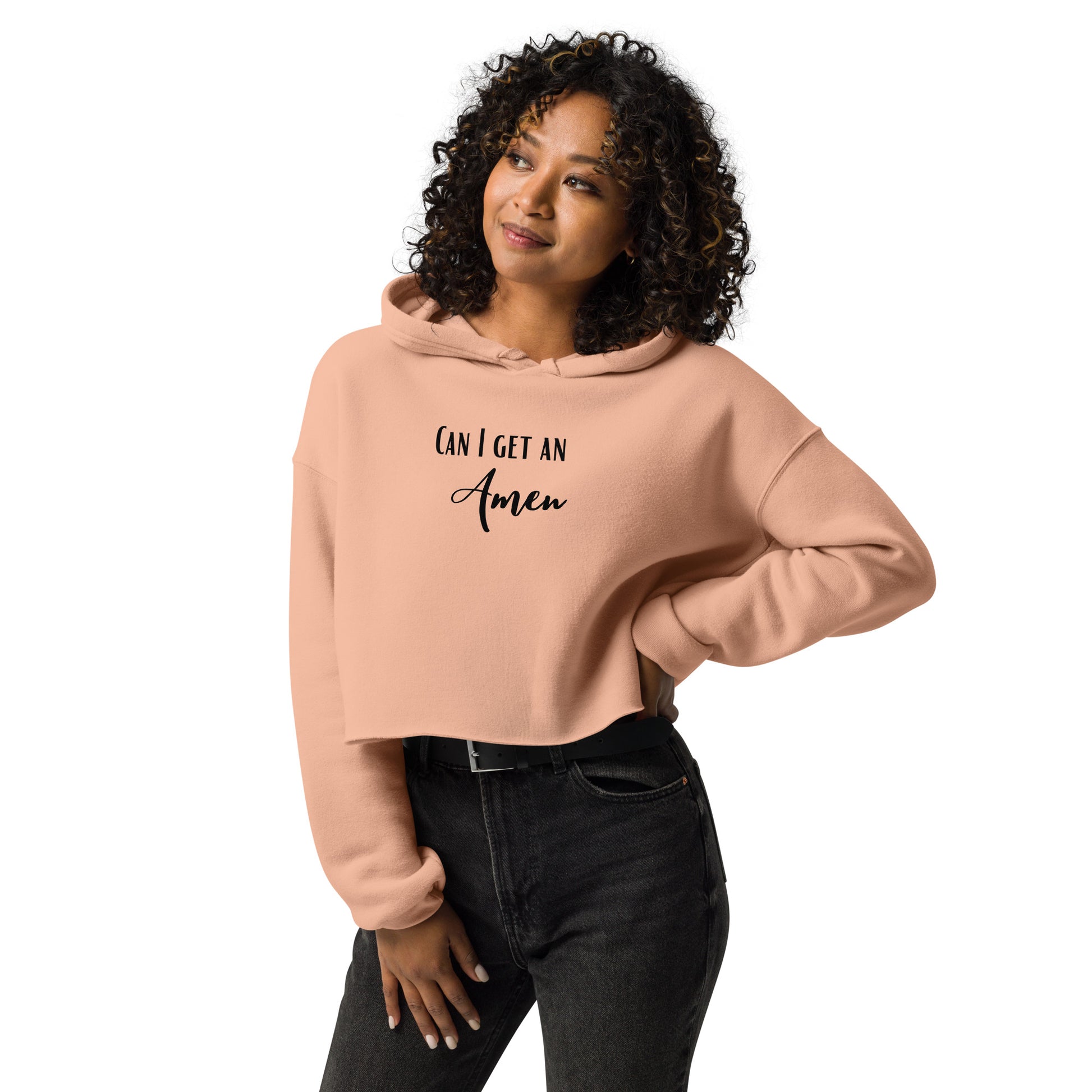 A pretty woman wearing a peach cropped hoodie or crop hoodie with long sleeves and the words Can I Get an Amen on the front in fun fonts, with black belted jeans, stands in front of a solid white background.