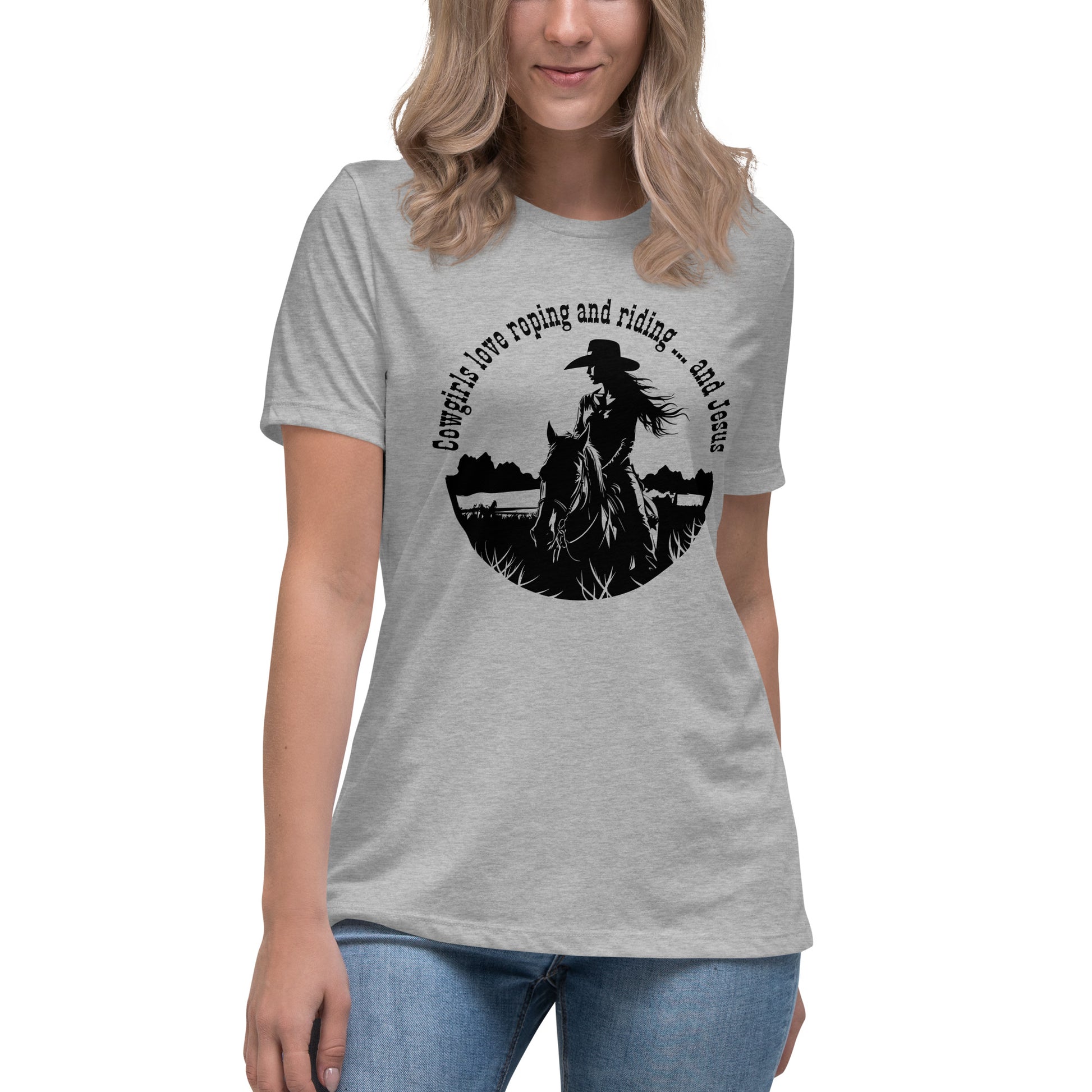 A front view of a woman from the nose to thighs wearing a light gray t-shirt with a design of a long-haired cowgirl on horseback and the words "Cowgirls love roping and riding ... and Jesus" all in a circular design.