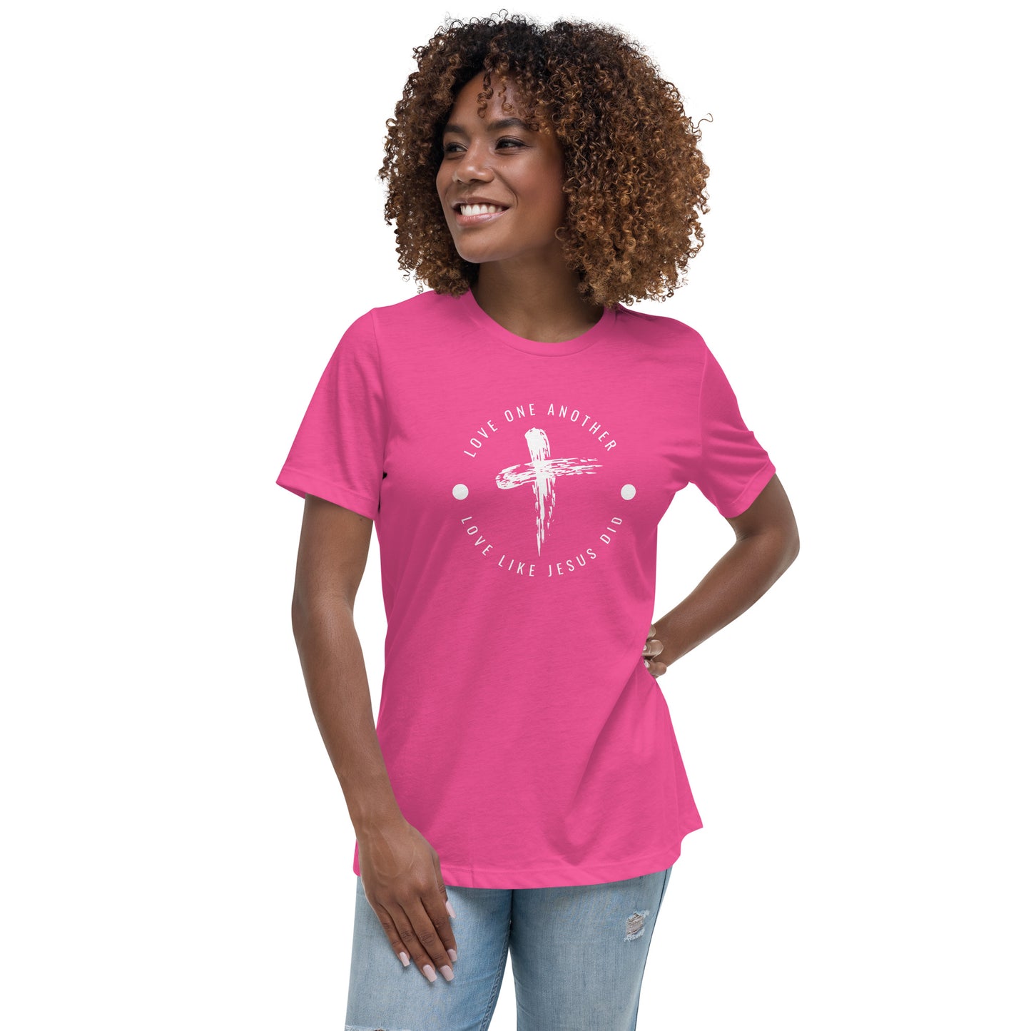 A smiling woman with short dark curly hair wearing light colored jeans and a bright pink short-sleeved t-shirt with a rustic cross in the center surrounded by words in a circle that say Love one another, Love like Jesus did.