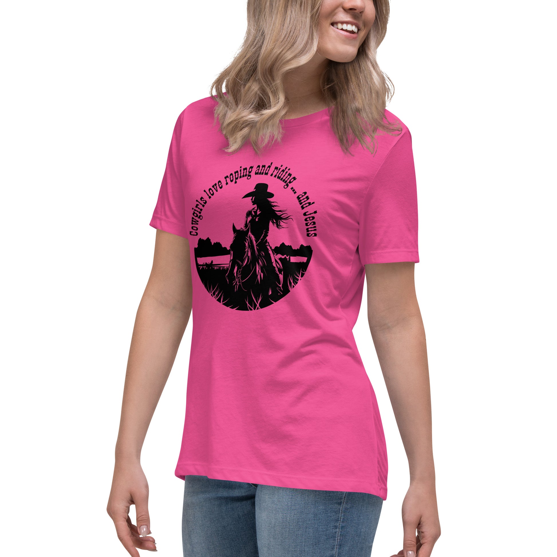 Partial view of a woman from nose to thighs and she's wearing a pink short-sleeved t-shirt and jeans. The t-shirt says Cowgirls love roping and riding ... and Jesus at the top and has a girl on horseback with a cowboy hat in a field cropped into a circle shape.