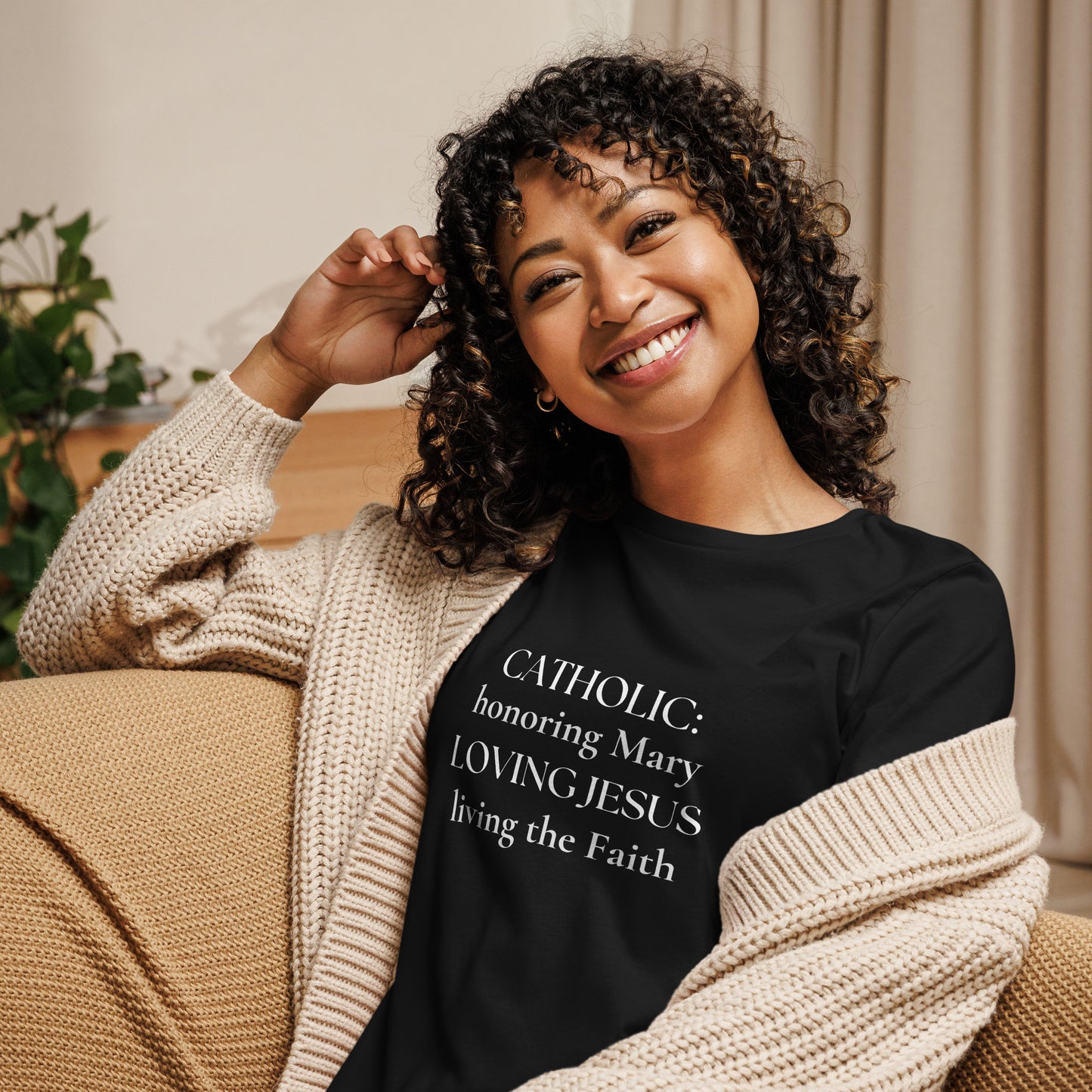 A smiling woman with curly hair is wearing a black t-shirt with white lettering that says:  CATHOLIC: honoring Mary LOVING JESUS living the Faith.