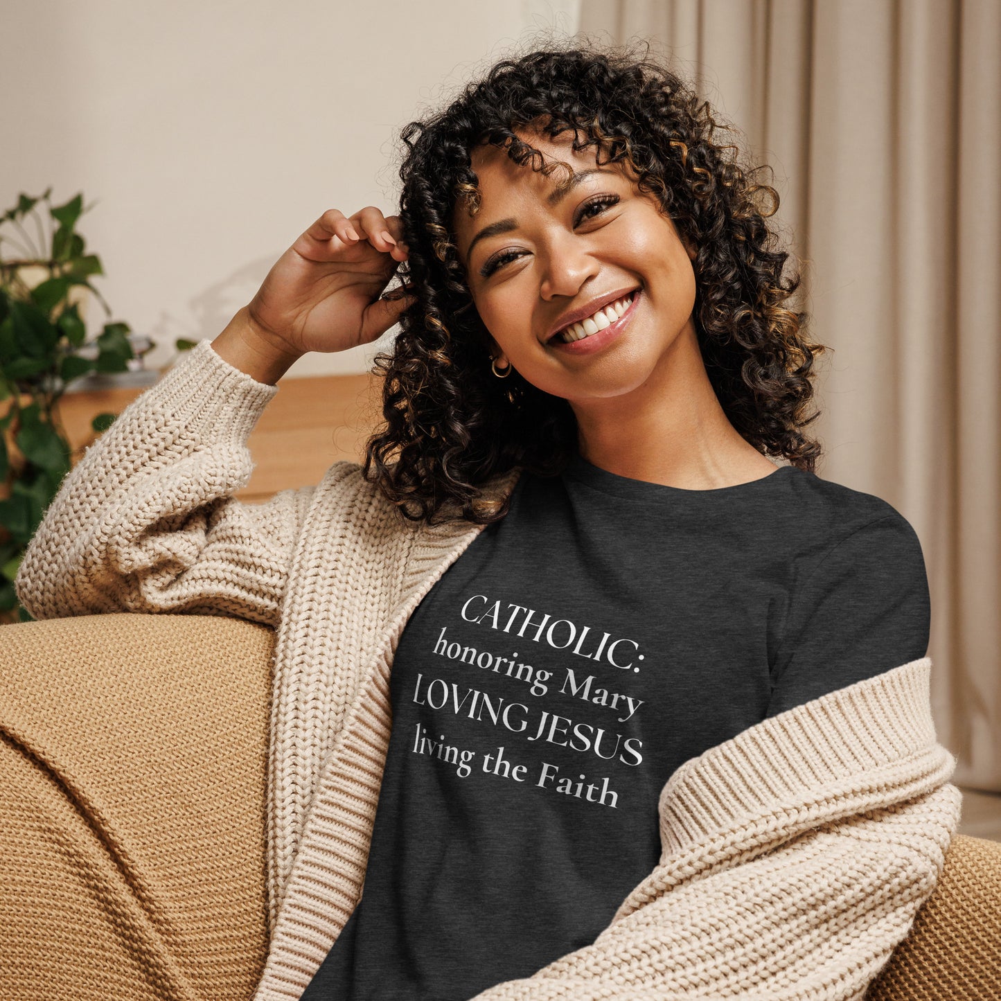 A dark heather gray t-shirt has white lettering that says:  CATHOLIC: honoring Mary, LOVING JESUS, living the Faith, and it is worn by a smiling woman with curly dark hair sitting on a sofa.