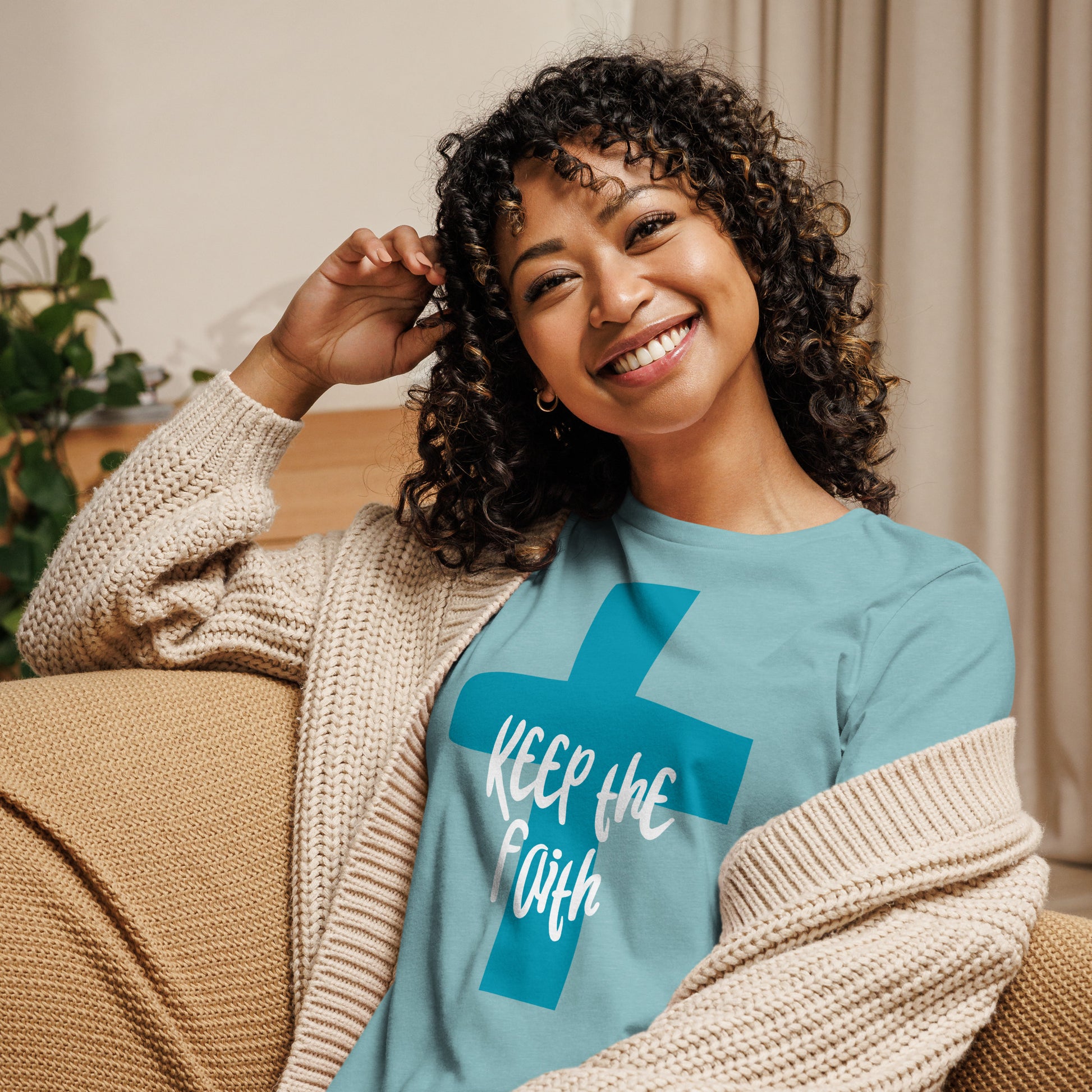 Seated woman on a couch, upper half of her body shown, with curly hair and a big smile, wearing an aqua t-shirt with a dark aqua cross and the words "Keep the Faith" in white on the cross, with a cream colored sweater slouched off one shoulder.