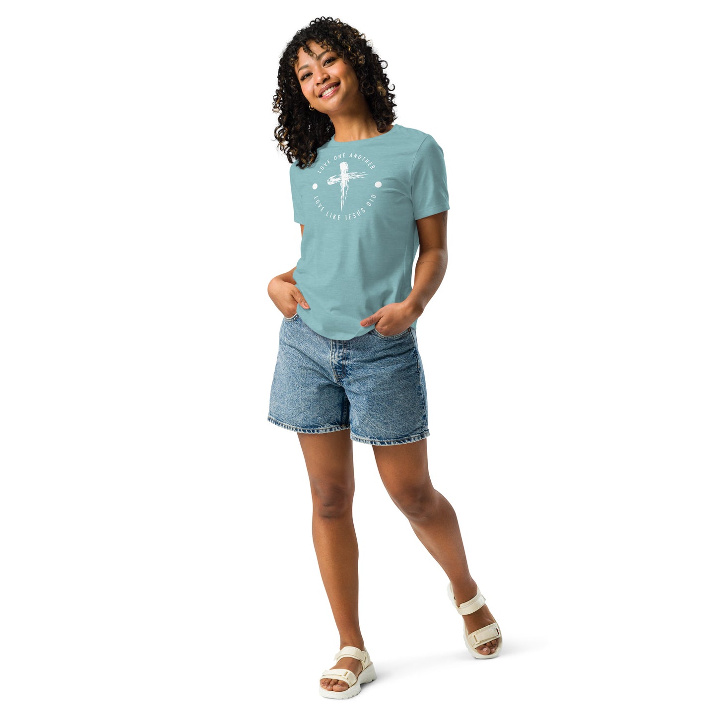 A young woman with dark curly hair, full length, wearing white sandals, long jean shorts, and an aqua t-shirt with words in a circle and a cross in the middle.