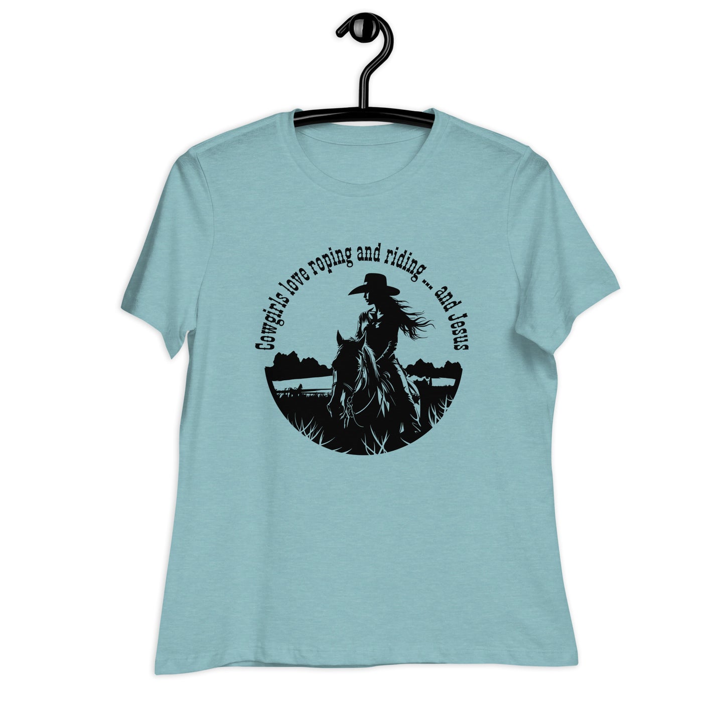 A solid white background with a light aqua t-shirt hanging on a black hanger which is hanging on a black hook. It features a cowgirl and horse design and the words:  Cowgirls love roping and riding ... and Jesus.