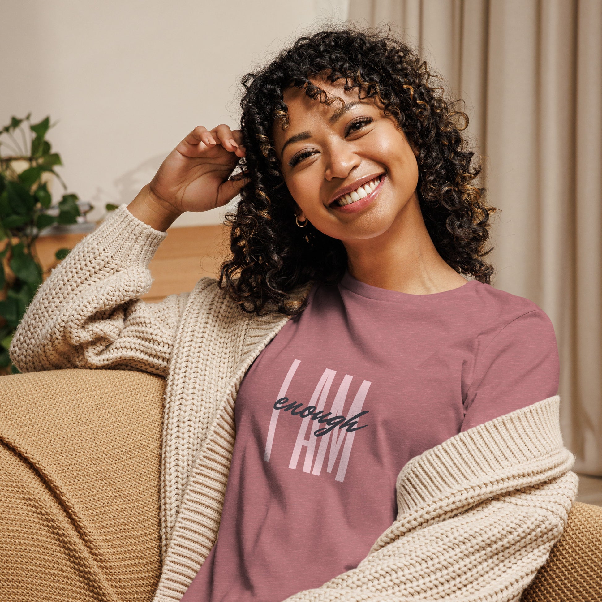 Smiling dark-curly-haired lady wearing mauve t-shirt with the words I AM enough in two different fonts and a cream colored cardigan over top, slid off left shoulder, relaxing n a couch.