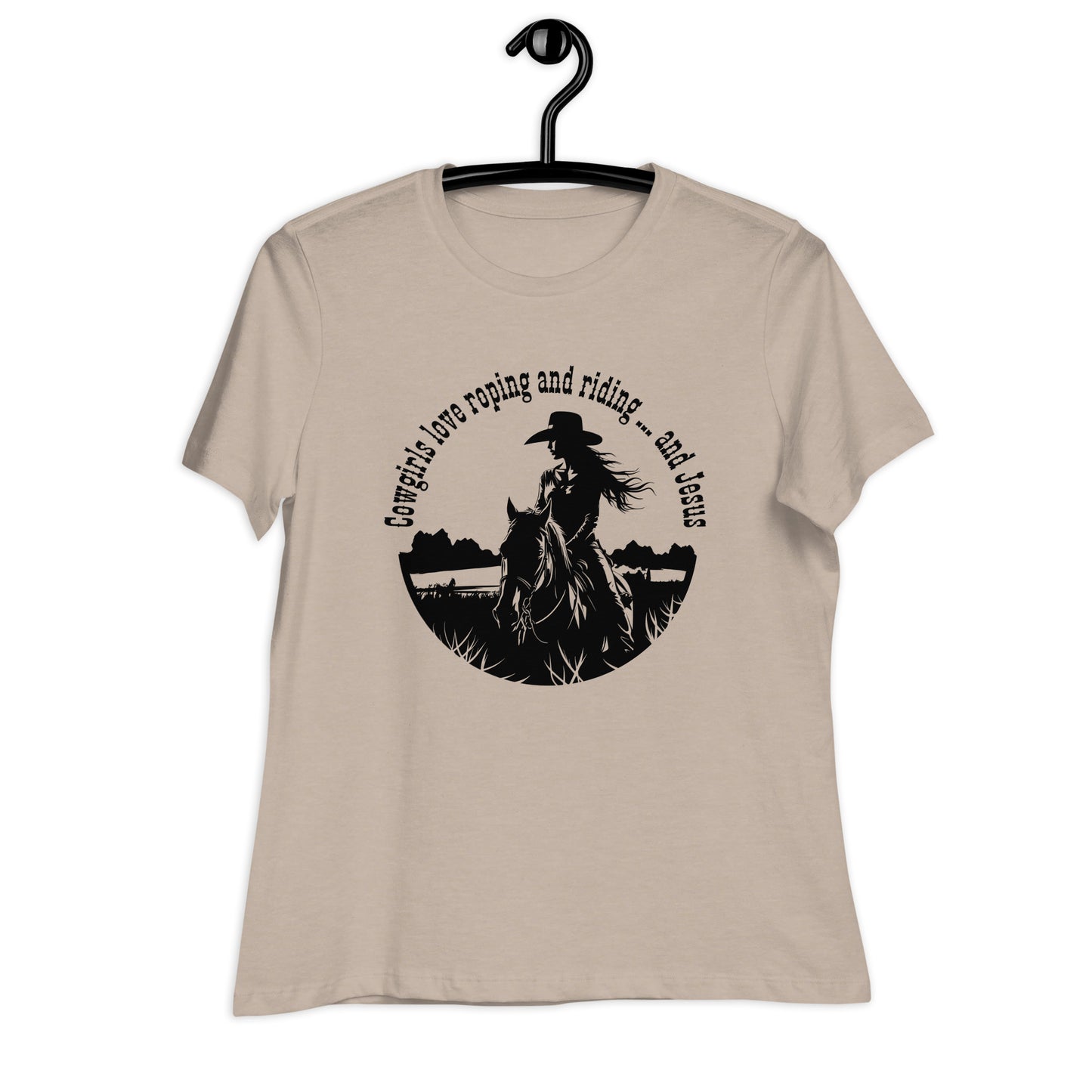 A tan t-shirt is hanging on a black hanger which hangs on a black hook. A black design on the front of the t-shirt shows a cowgirl on a horse in a field or pasture and says:  Cowgirls love roping and riding ... and Jesus.