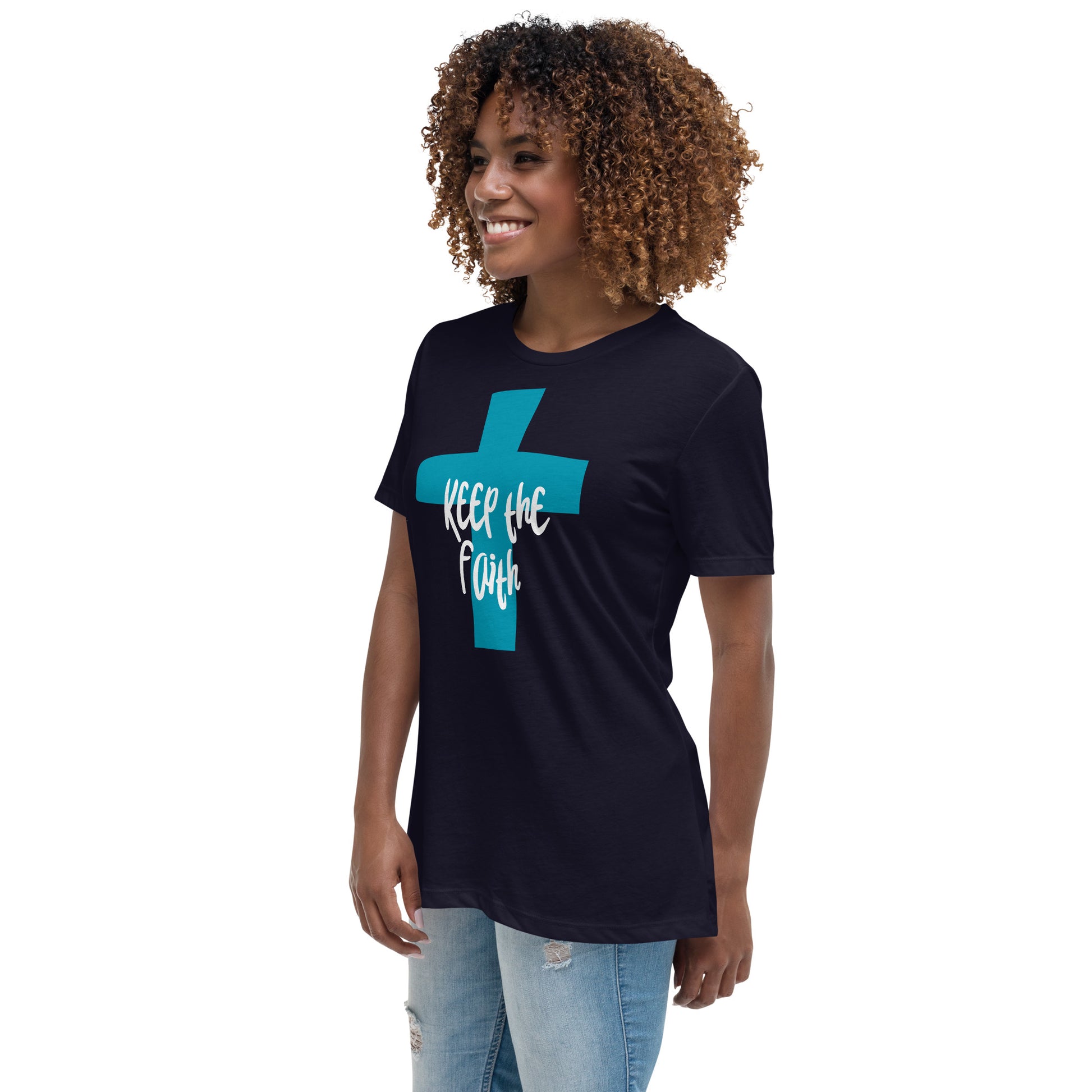 Three-quarter length view of a smiling woman with curly hair wearing light denim distressed jeans and a black short-sleeve t-shirt with a bold dark aqua cross on the front, and over part of the cross in large white letters it says Keep the Faith in a cool font.