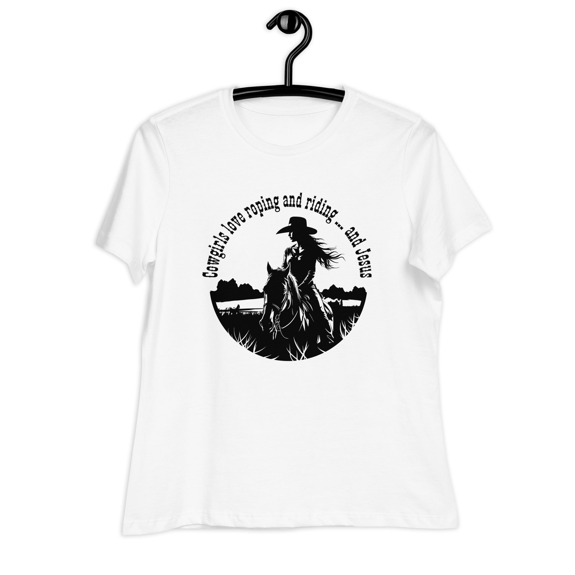 A white t-shirt on a black hanger features a black circular design on the front of the short-sleeved t-shirt. The design says Cowgirls love roping and riding ... and Jesus in an arc at the top of a circle and then there is a cowgirl on horseback making up the rest of the circular design.